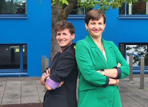Freddie* and Simone are standing in front of a tree with their backs to each other and their arms crossed. Both are wearing blazers and grinning at the camera