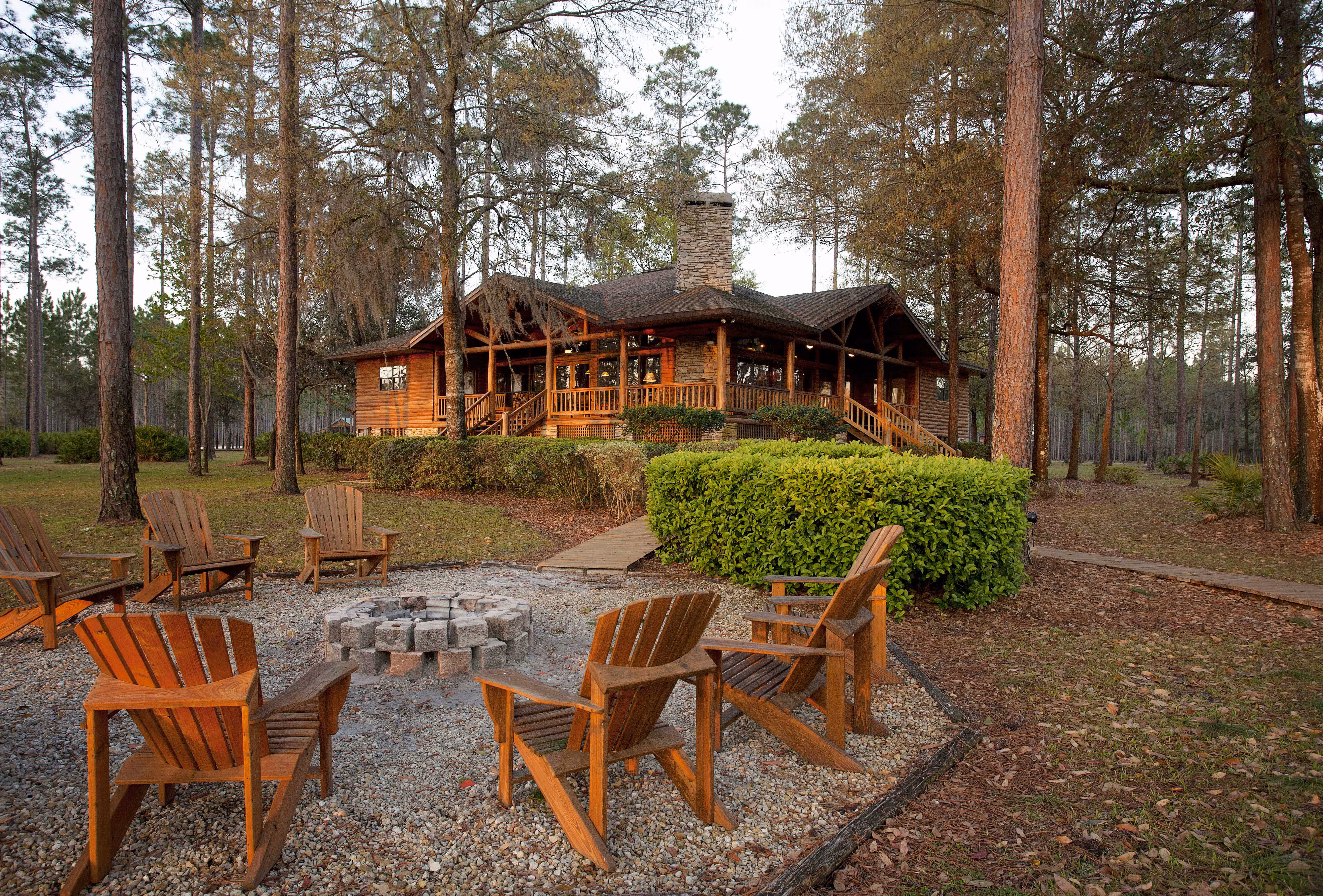 Wooden Adirondack chairs arranged around a stone fire pit in front of a rustic cabin surrounded by tall trees.
