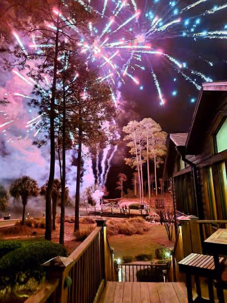 Colorful fireworks burst in a night sky above tall trees and a wooden deck with chairs near a building.