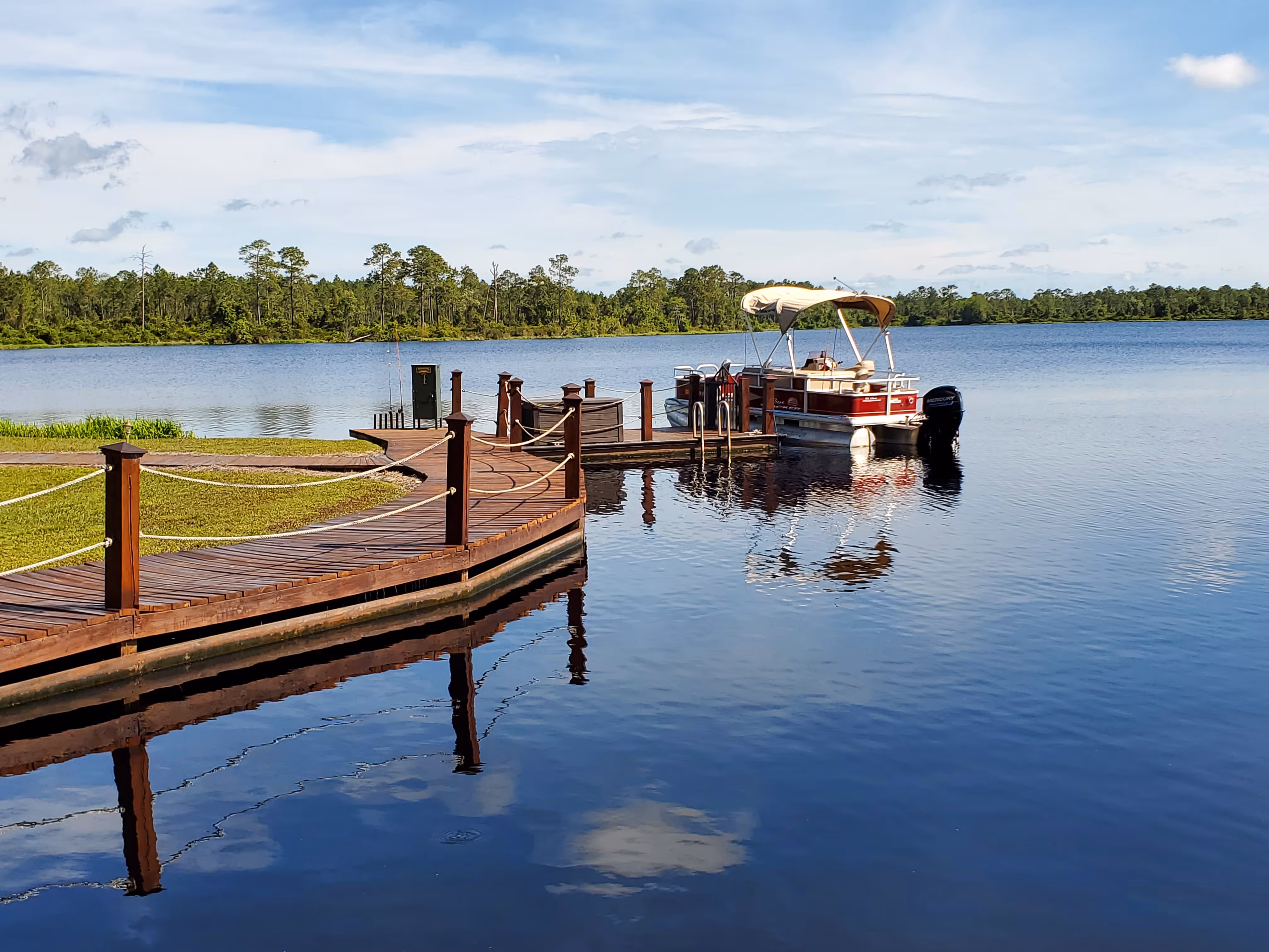 Wooden dock with rope railings extending over a calm lake with a pontoon boat moored at the end.