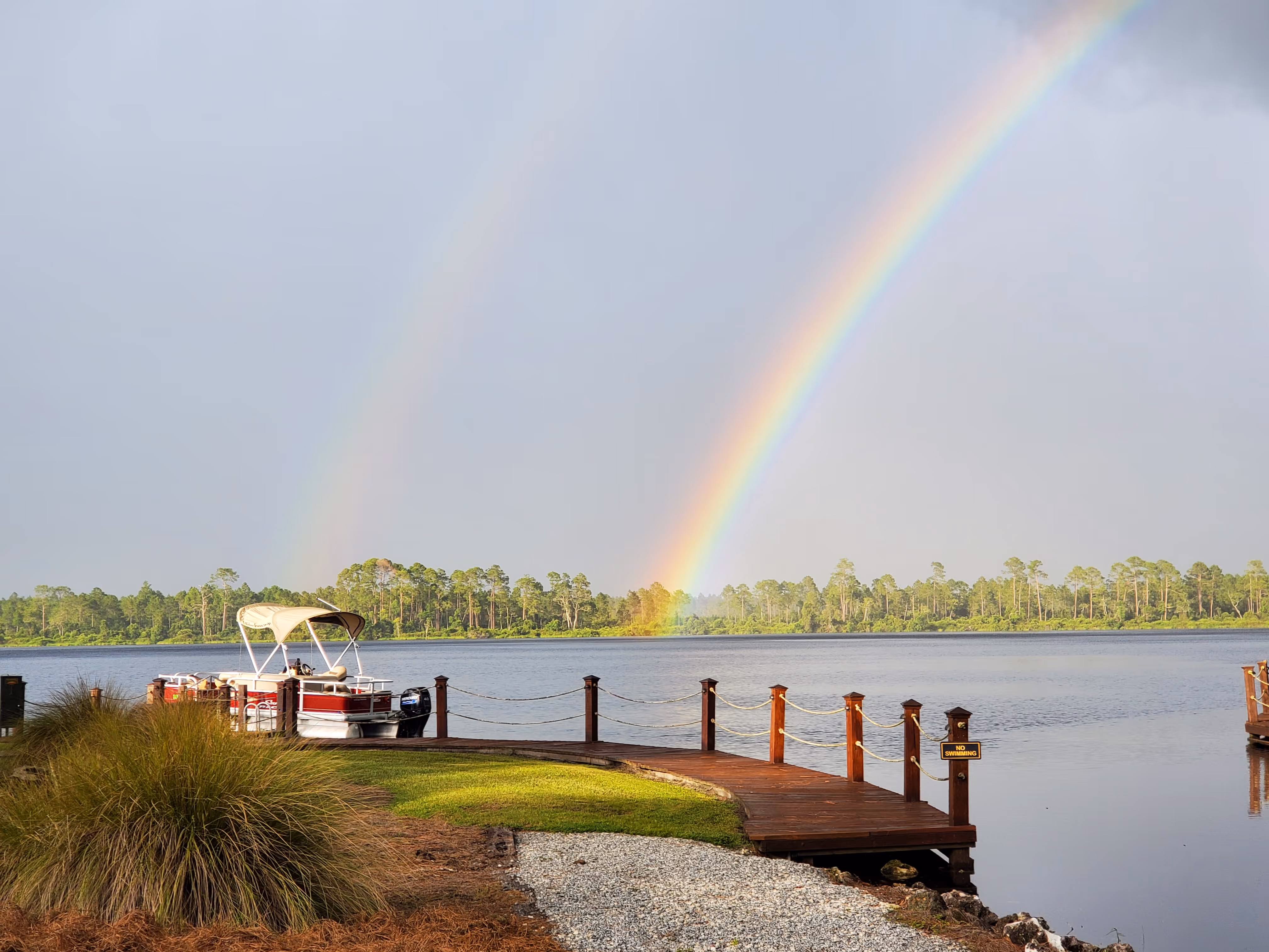 Double rainbow over a calm lake with a dock and a moored pontoon boat on the left side.