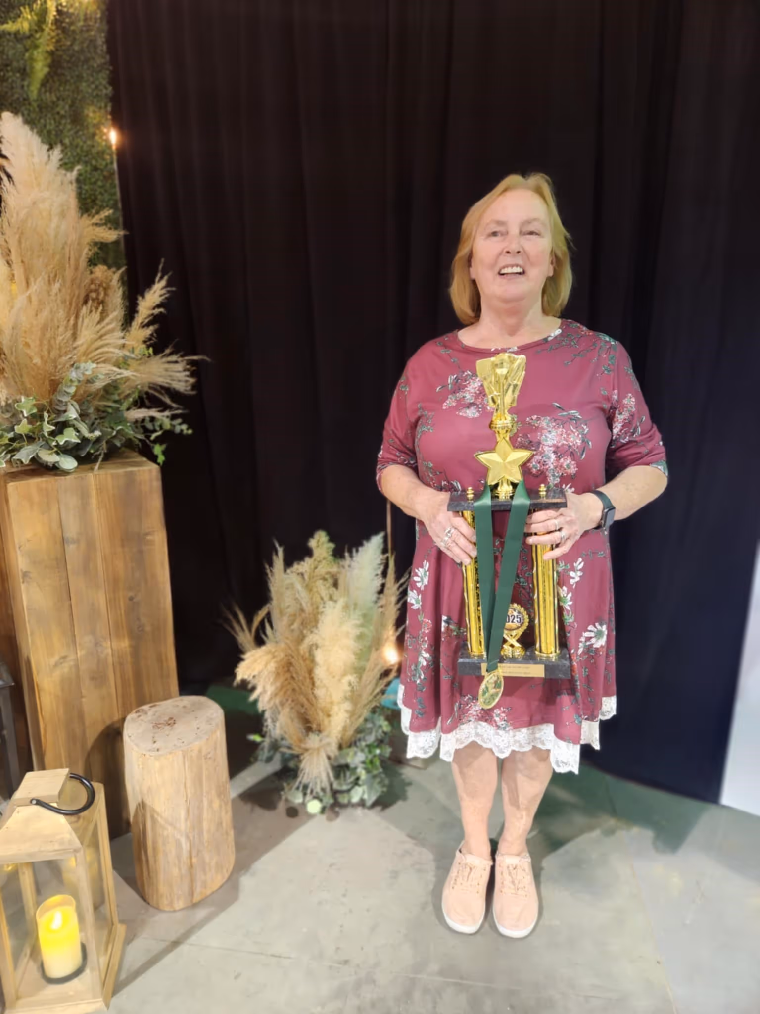 Woman in a floral maroon dress holding a large gold trophy with a green medal ribbon, standing indoors near wooden decorations and dried plants.