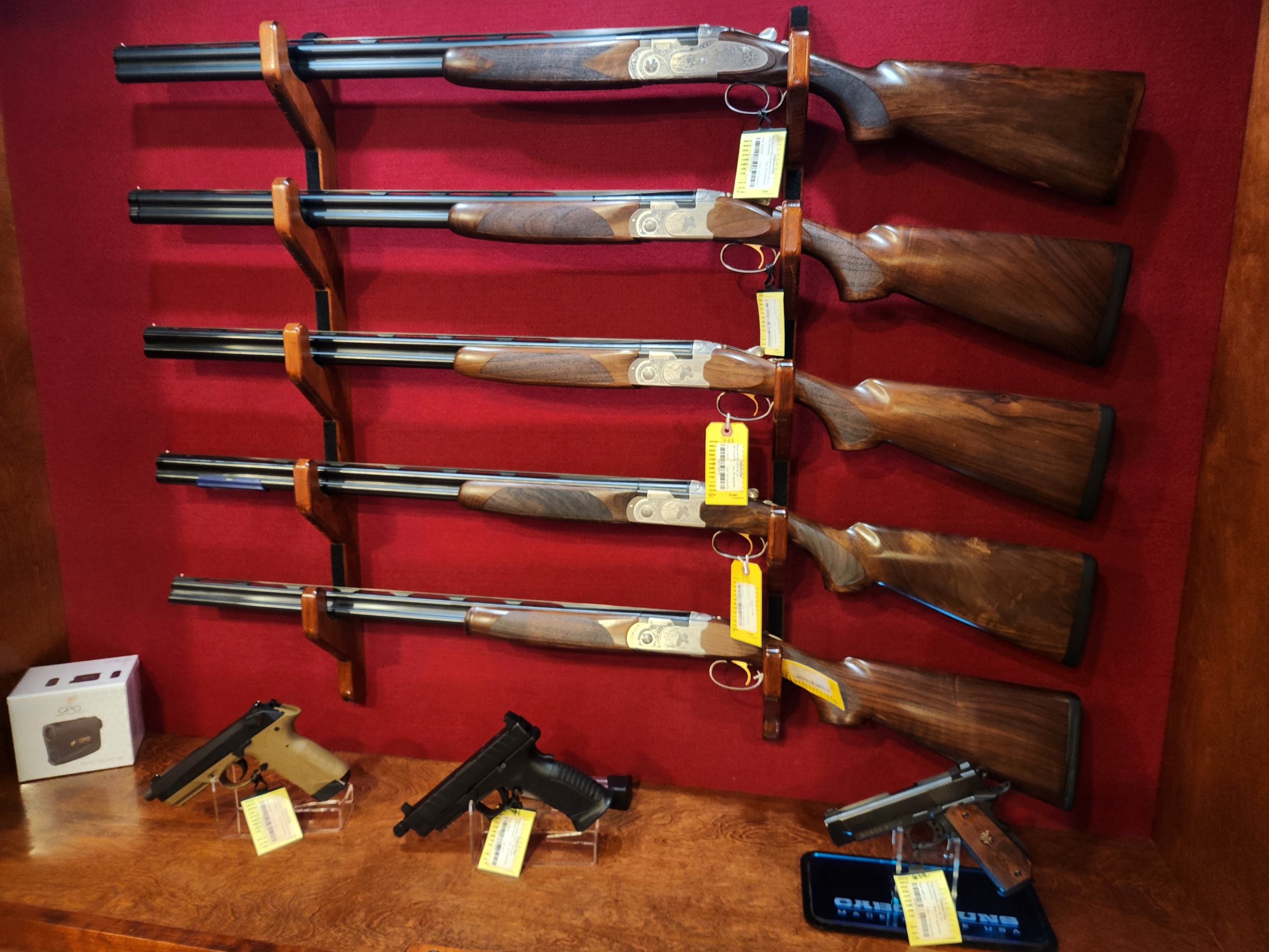 Five double-barrel shotguns mounted vertically on a red wall above three handguns displayed on a wooden surface, all with price tags.