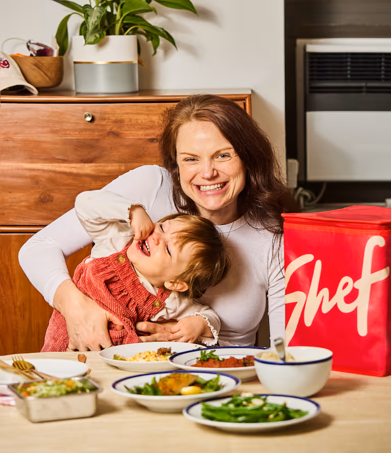 Smiling woman hugging a laughing toddler at a dining table with multiple dishes and a red insulated bag labeled 'Shef'.