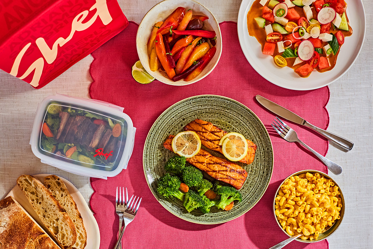 Grilled salmon with lemon slices and steamed broccoli on a plate, surrounded by bowls of macaroni and cheese, roasted carrots, a fresh salad, sliced bread, and a container of steak salad on a red placemat.