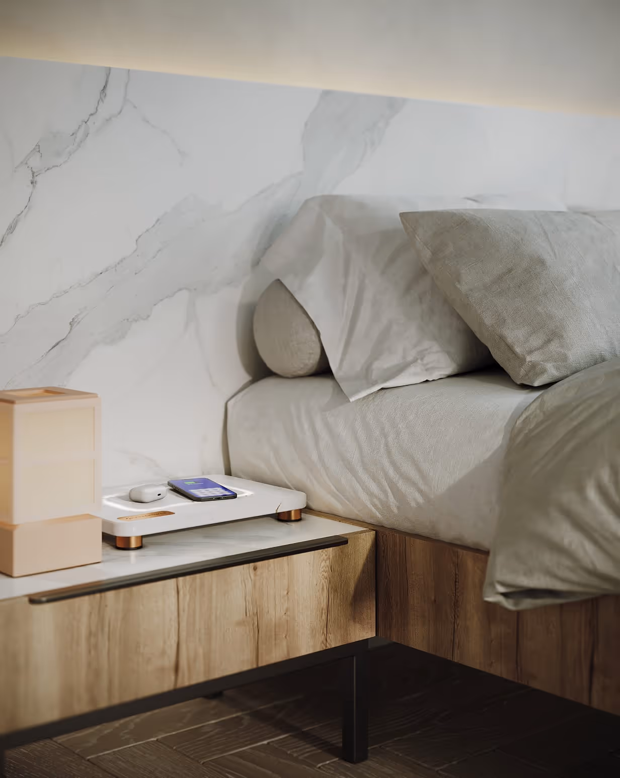 Modern bedside table with wooden drawer, white FreePower Valet wireless charging device with phone and earbuds, next to a bed with gray pillows and light gray bedding.