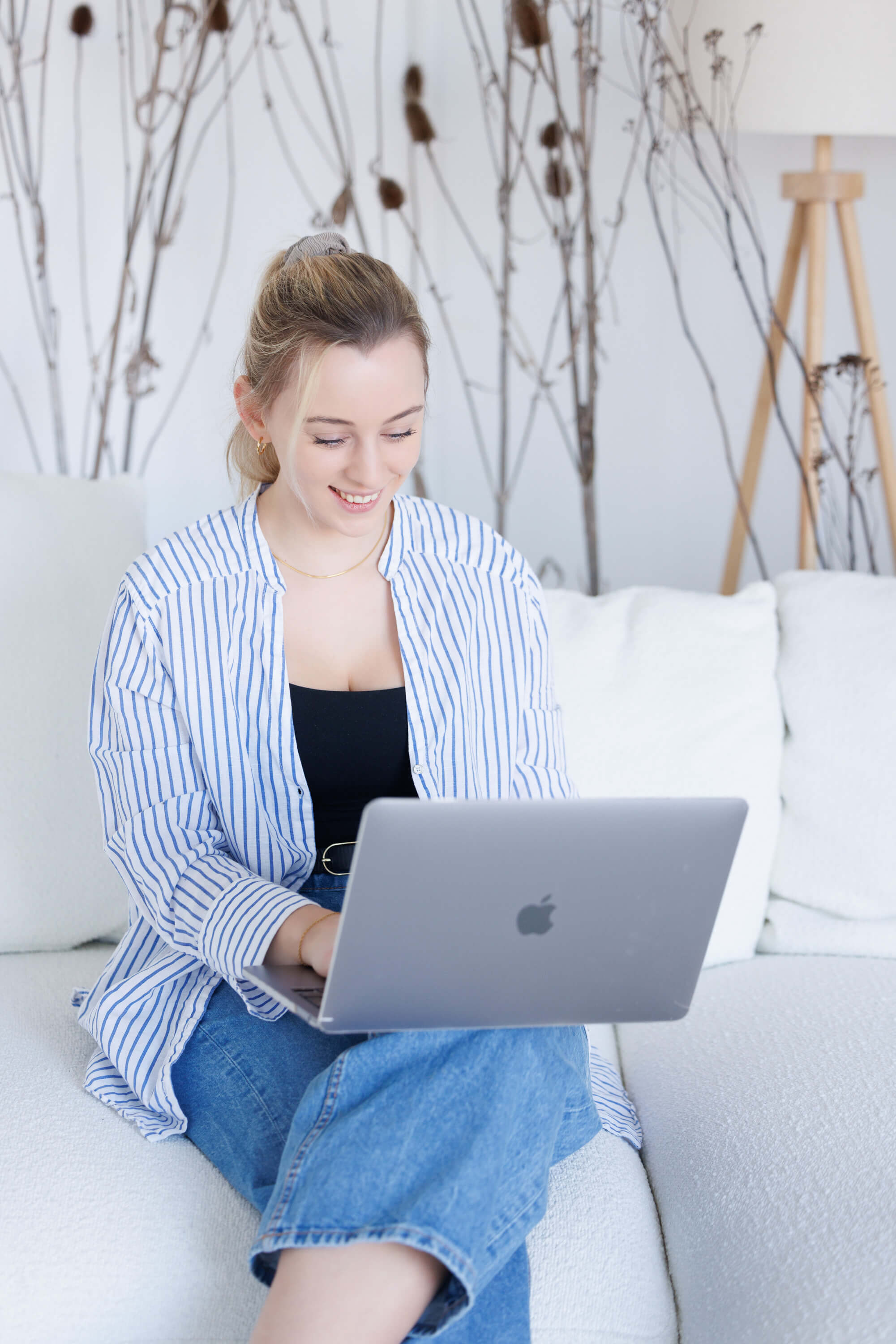 Tina Hörholdt in gestreifter Bluse lächelt über ihren Laptop, auf einem weißen Sofa