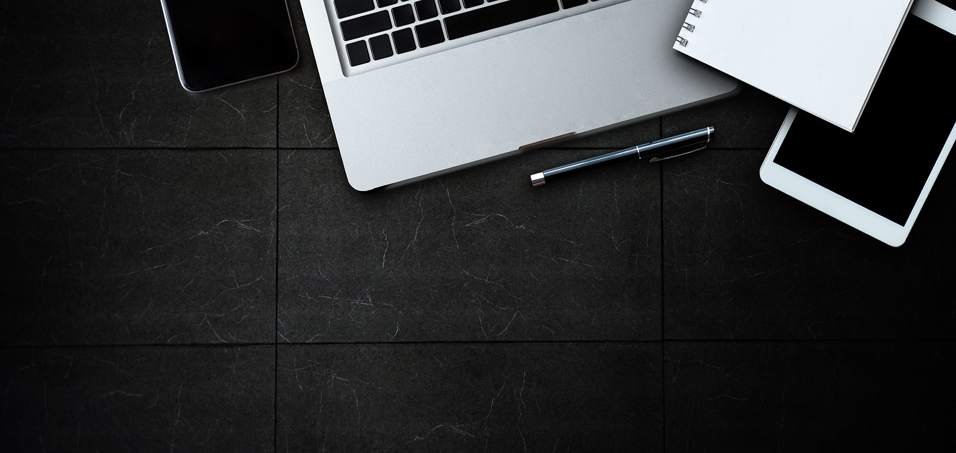 Top view of a dark office desk with a laptop, smartphone, tablet, pen, and a blank notebook.