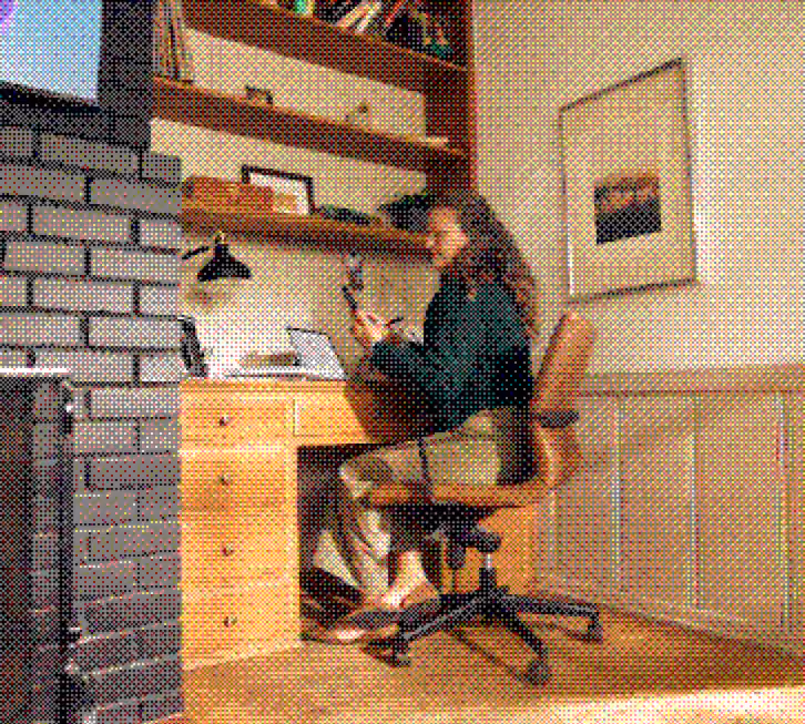 Woman sitting on a swivel chair, working at a wooden desk in a cozy room with bookshelves and a framed picture.