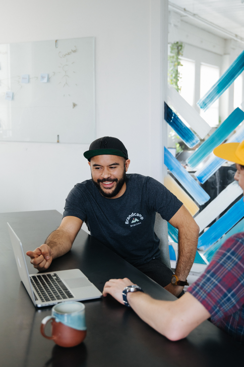 Four young professionals gathered around a table in a modern office, smiling and looking at laptops.
