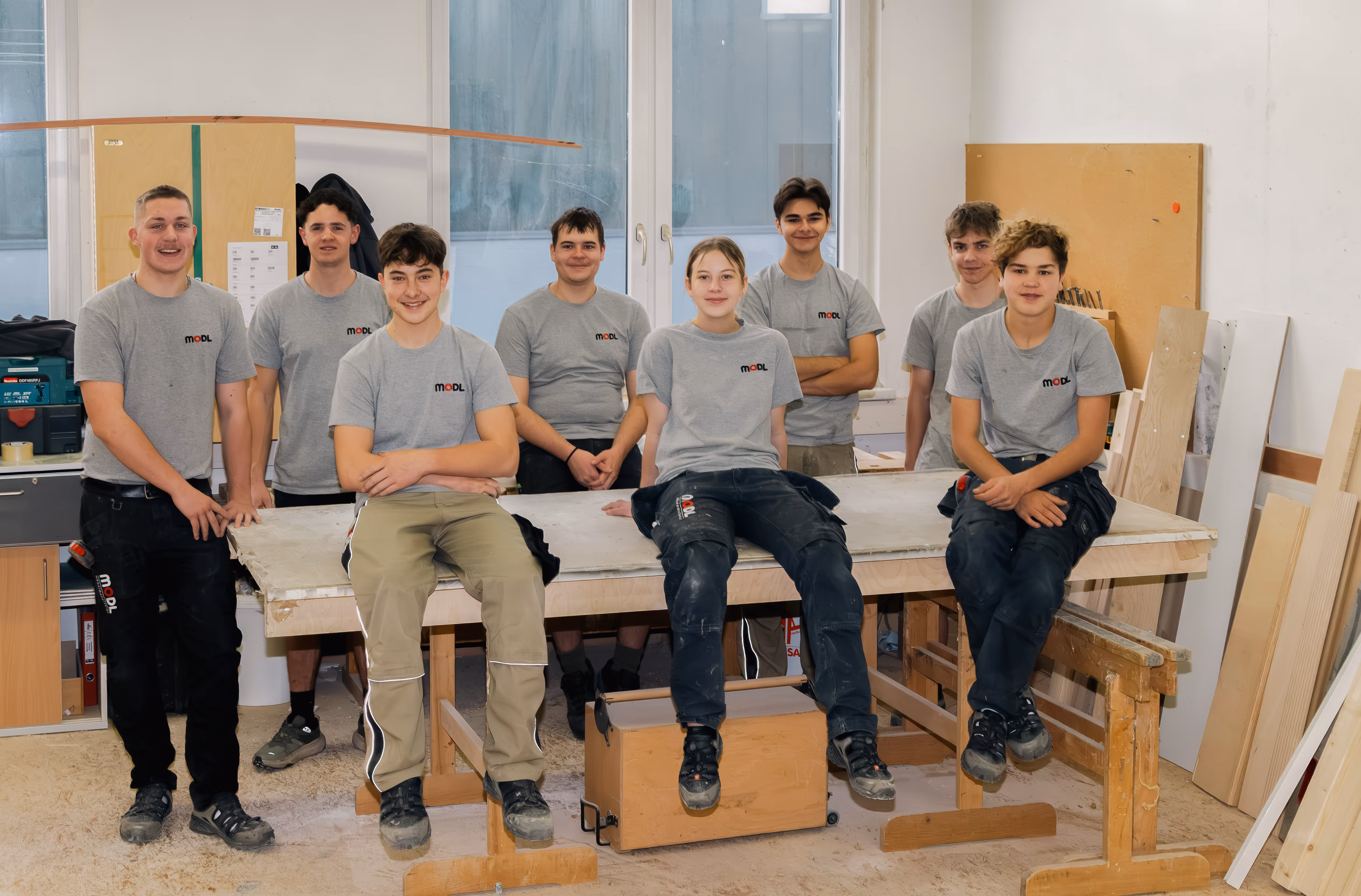 Acht junge Handwerker in grauen Modl-T-Shirts posieren in einer Werkstatt, einige sitzen auf einem Arbeitstisch, andere stehen dahinter.