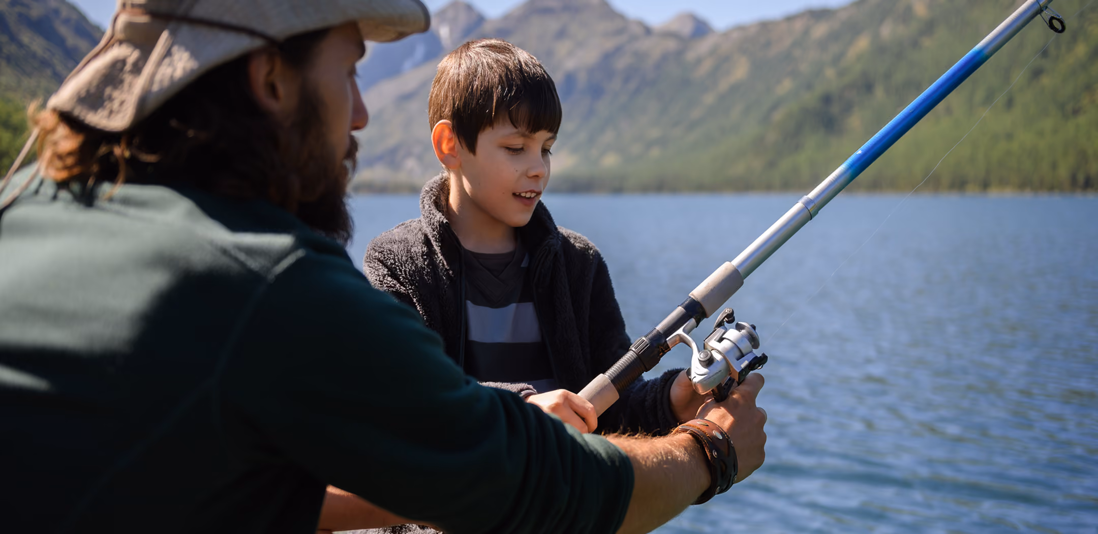 Man helping a boy hold a fishing rod beside a lake