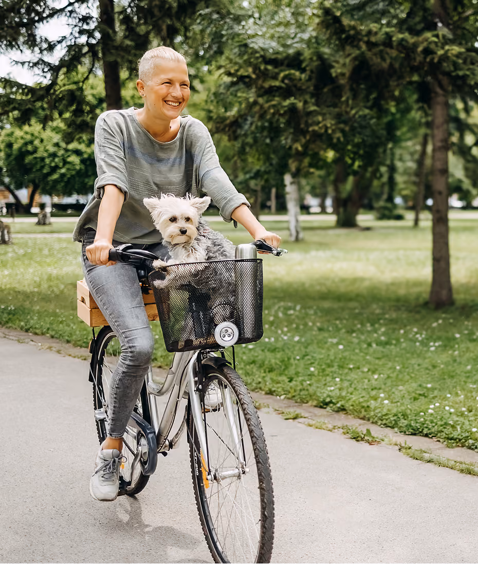 Woman cycling through a park with a small dog in the front basket