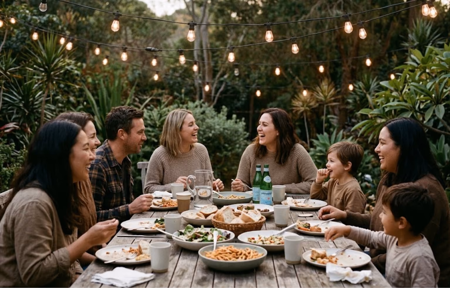 Adults and children eating together at a long outdoor table under string lights