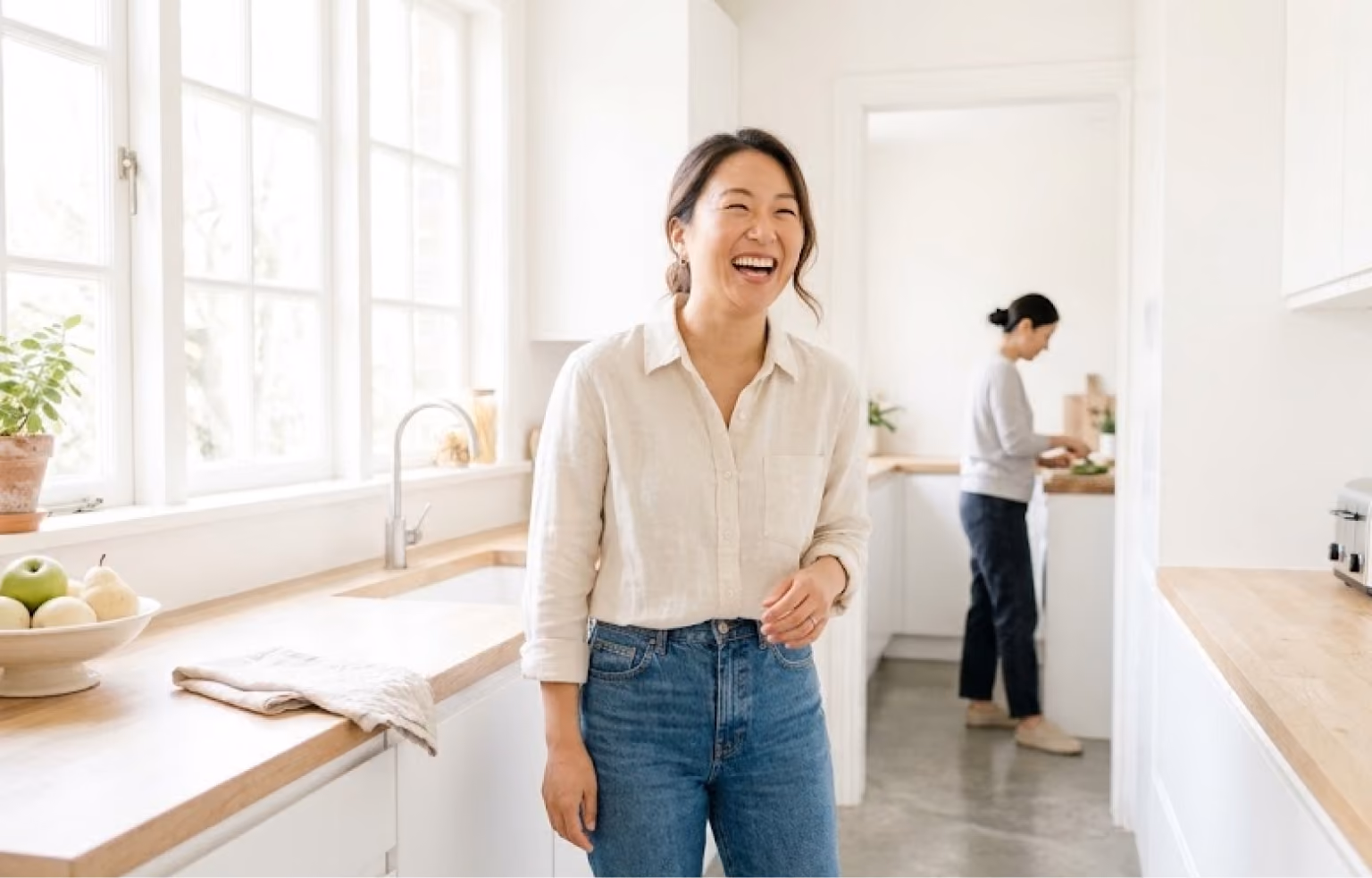 Woman laughing in a bright kitchen while someone works behind her