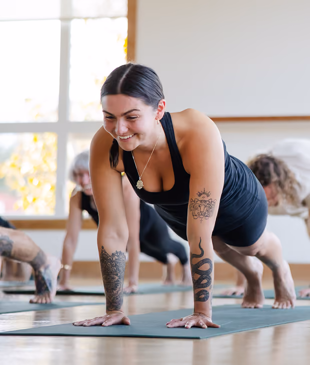 Young woman with tattoos on her arms smiling while holding a plank yoga pose on a mat in a bright studio with others in the background.