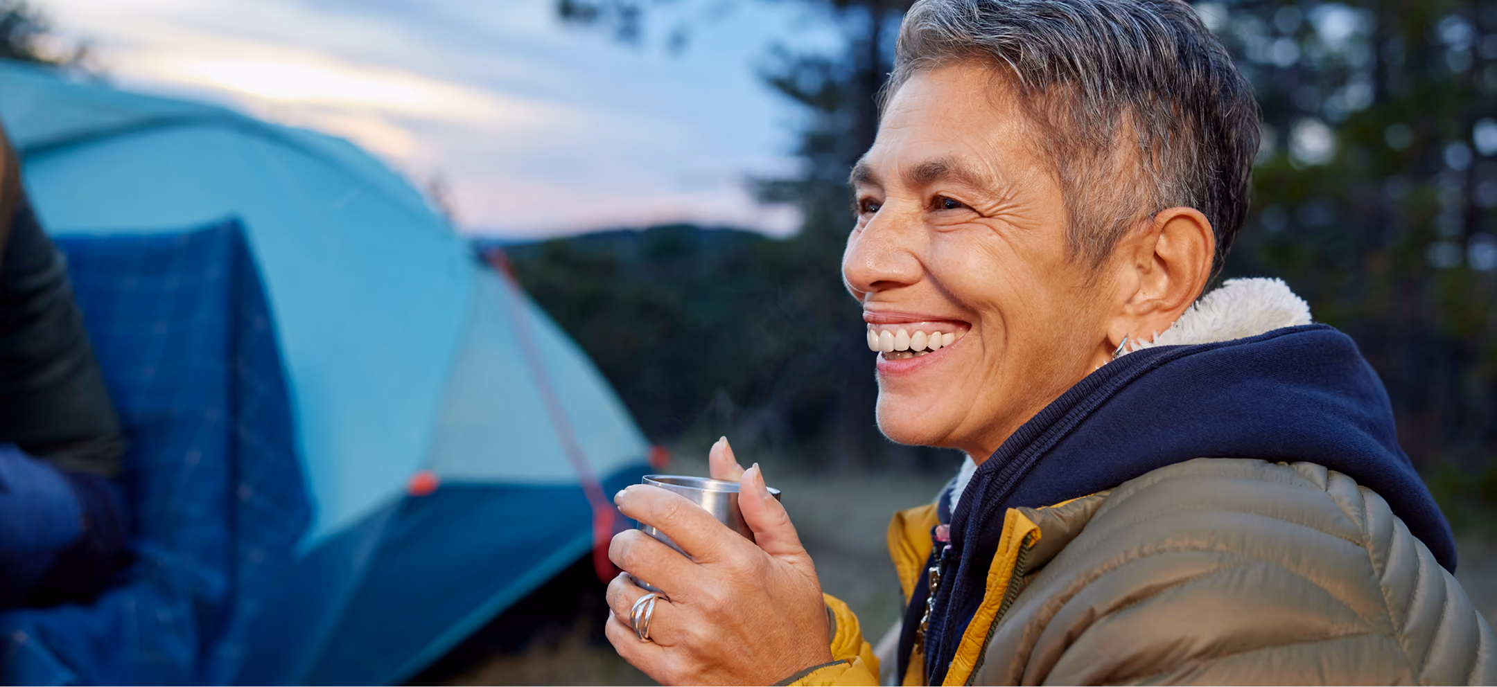 Smiling person wearing a yellow and green jacket holding a metal cup, sitting outdoors near a blue tent during sunset.