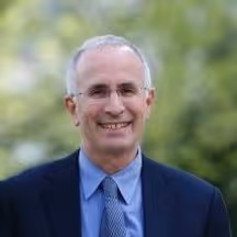 Professional photograph of Russ Phillips, an older man with short grey hair and glasses, wearing a dark suit, blue shirt and striped tie, smiling outdoors with green foliage blurred in the background.