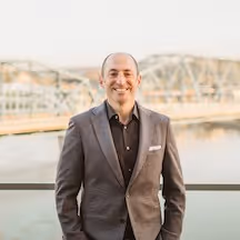Professional photograph of Benjamin Miller, a bald man with a trimmed beard, wearing a grey checked blazer over a black shirt, smiling outdoors with a bridge and river visible in the background.
