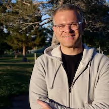 Professional photograph of Bryan J Hampton, a man with short fair hair, wearing rimless glasses, a light grey zip up hoodie and a black shirt, smiling outdoors with arms crossed and trees visible in the background.