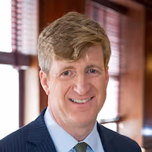 Professional headshot photograph of Patrick Kennedy, a man with short fair hair, wearing a pinstripe dark suit, light blue shirt and yellow tie, smiling against an office background with window blinds.