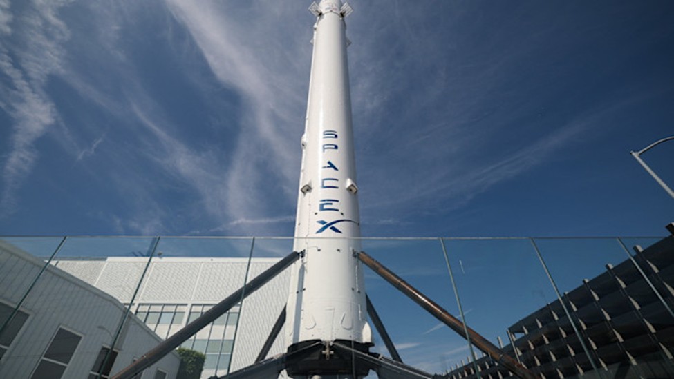 A vertical SpaceX Falcon 9 rocket booster standing outdoors against a blue sky with wispy clouds.