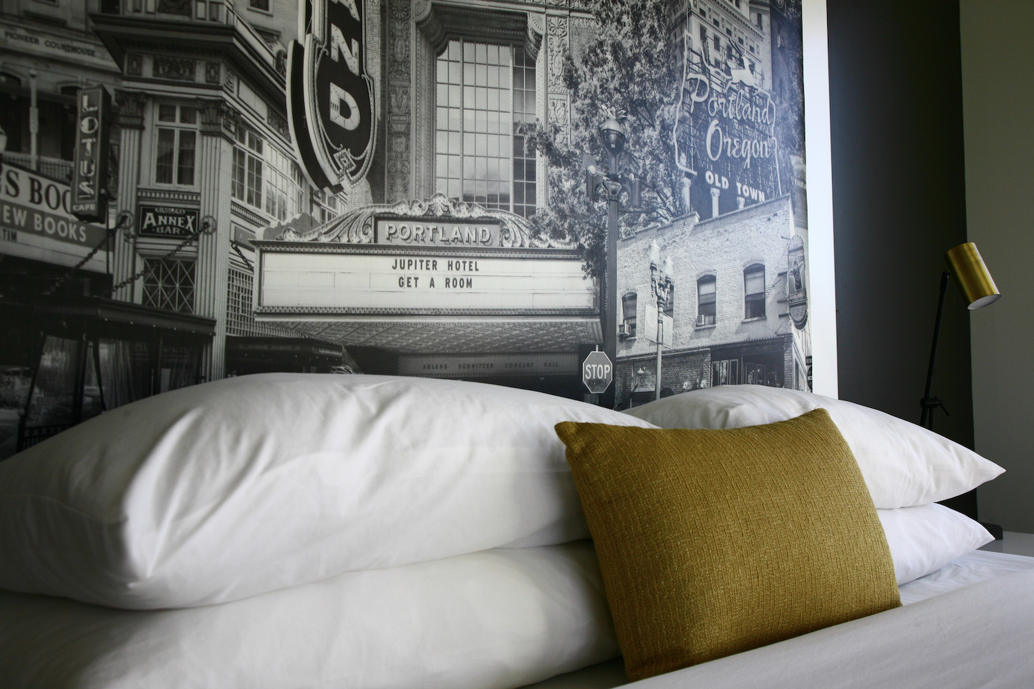 Bed with white pillows and a mustard-yellow pillow in front of a black-and-white mural of Portland's old town featuring a theater marquee reading 'Jupiter Hotel Get A Room.'