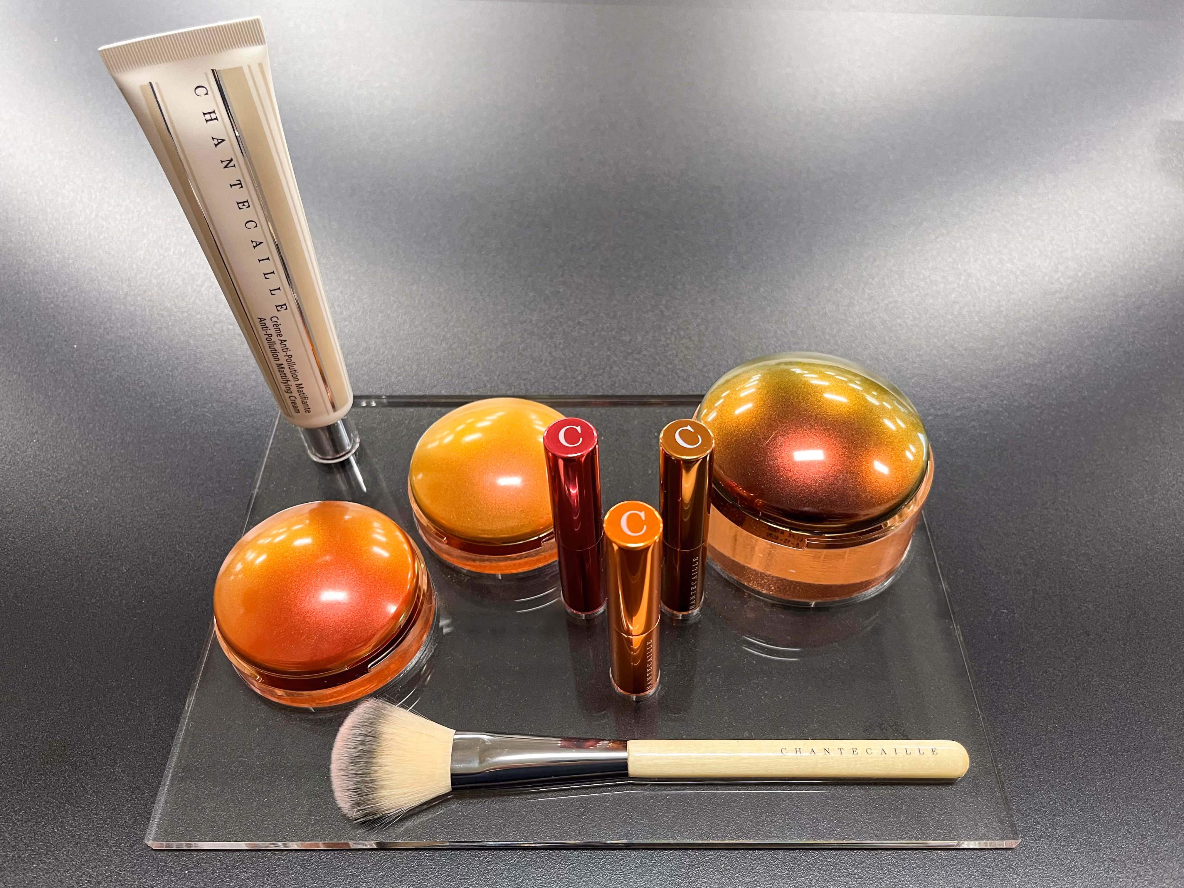 Chantecaille makeup products including tubes, jars, lipsticks, and a brush on a clear display stand.