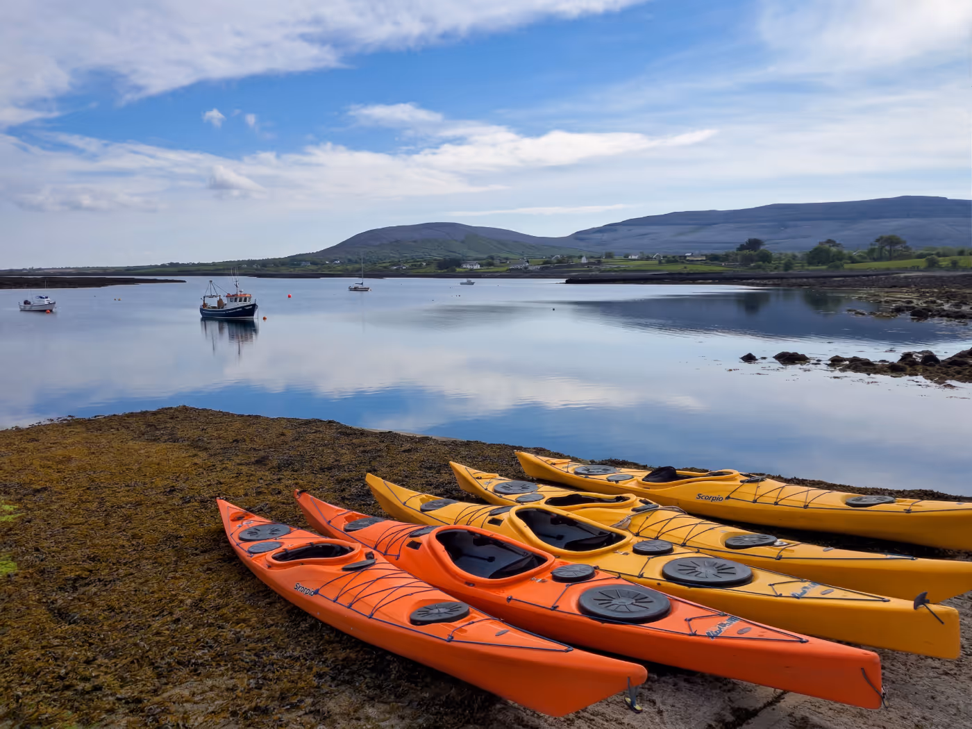 Orange and yellow kayaks on shore with calm water, boats, hills, and cloudy sky in background.