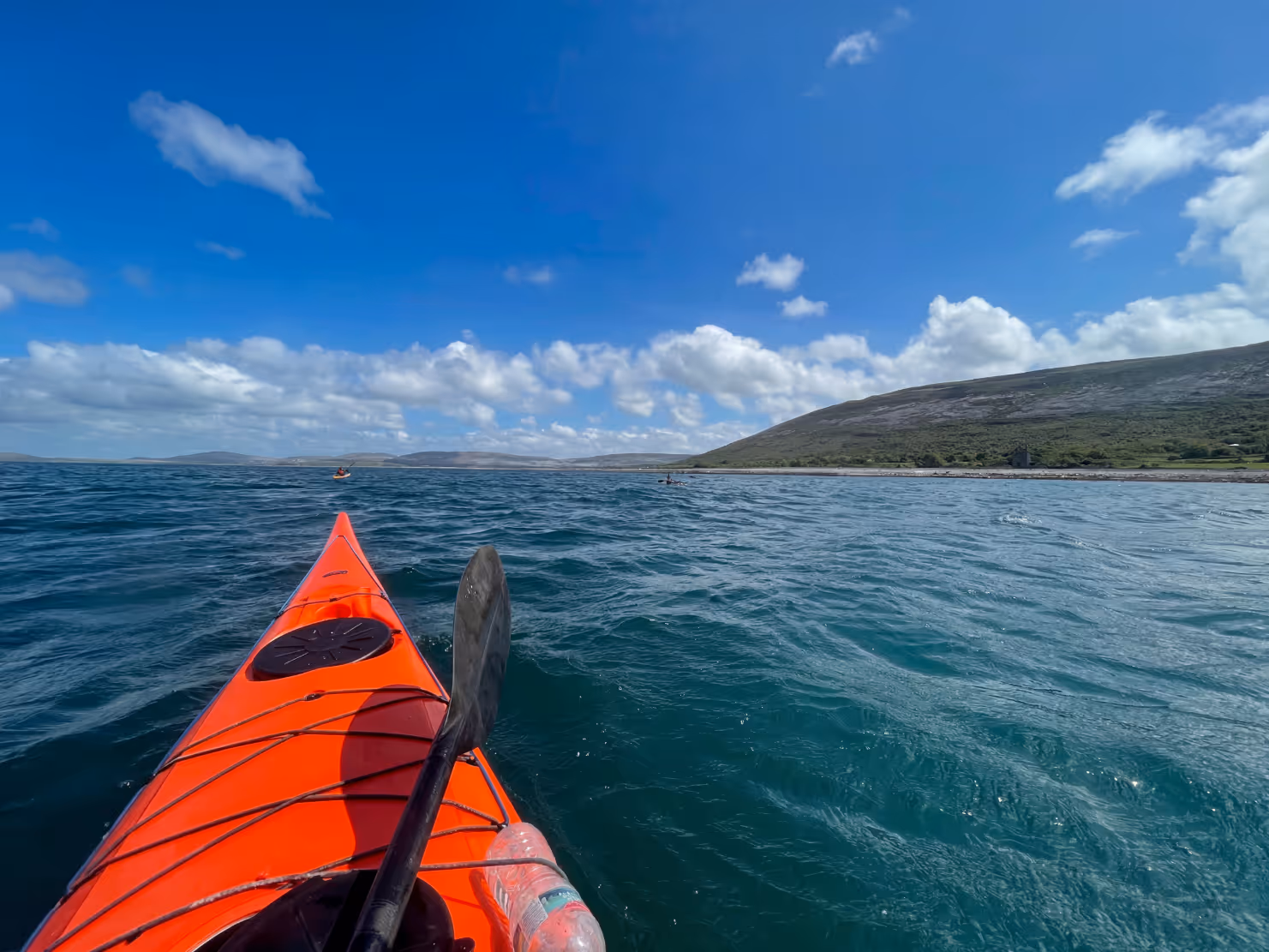 View from a red kayak paddling on blue water under a partly cloudy sky near a rocky shoreline.