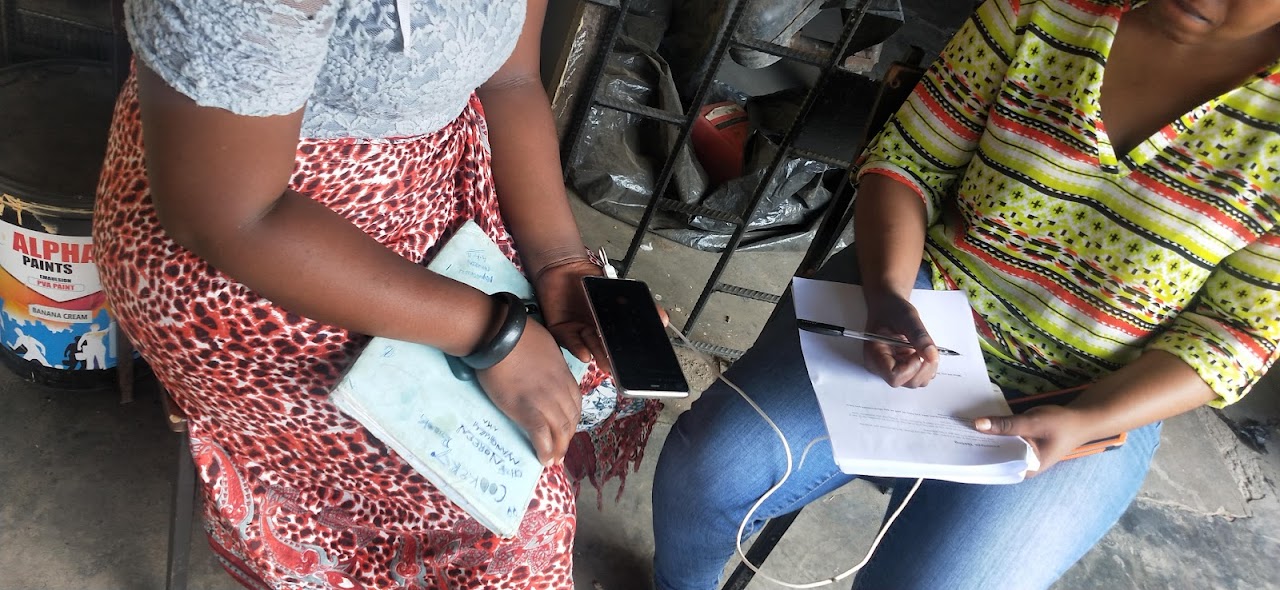 Close-up photograph showing the torso of two women in Zambia talking to each other, one is holding a phone, the other writing notes on paper