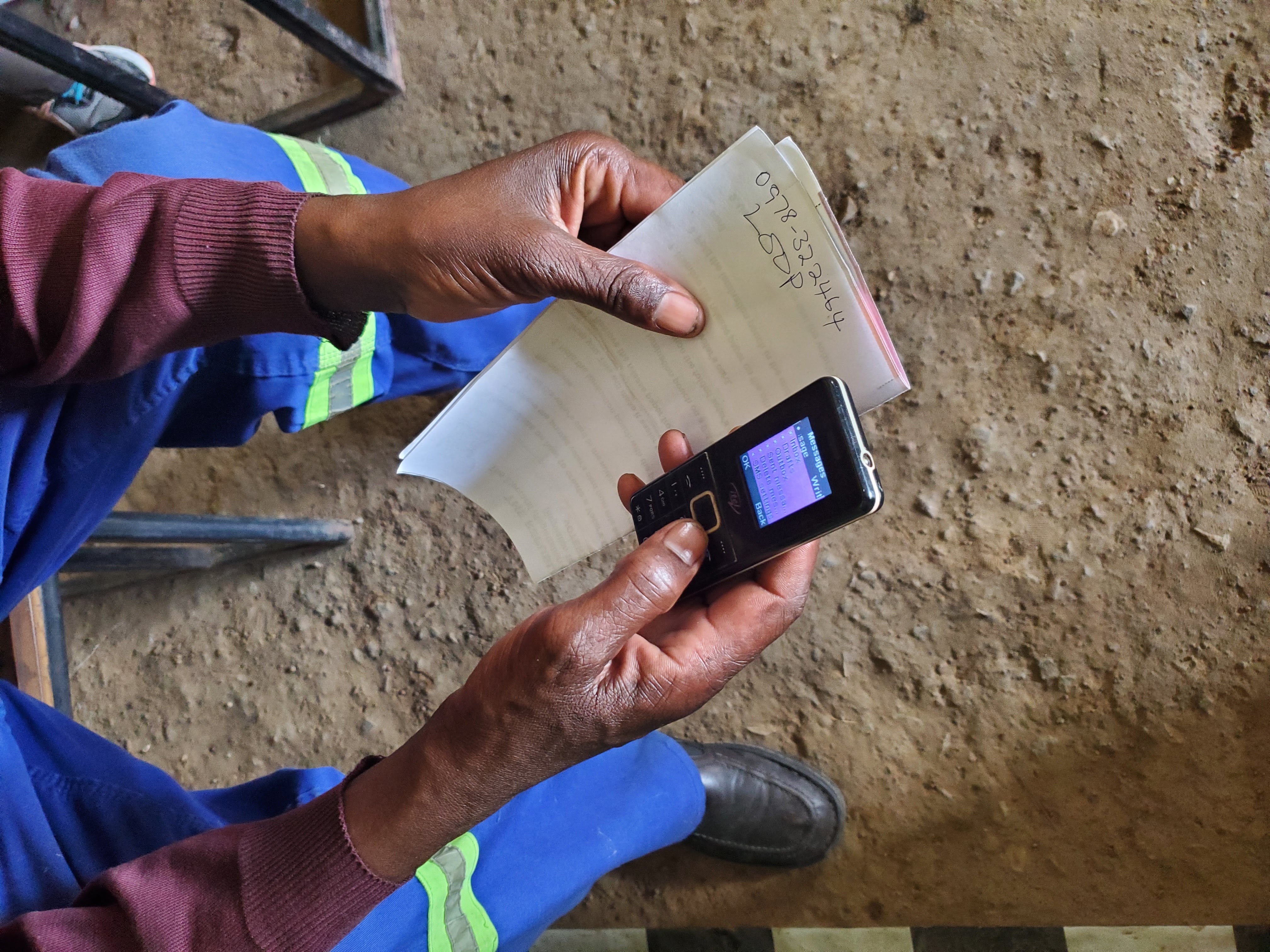 A man holds a simple mobile phone and a piece of paper as he writes a text message to talktoloop.org in Zambia