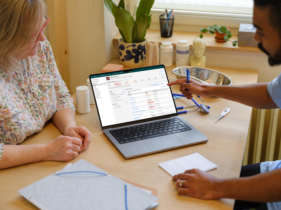 A healthcare worker points to a medical health record displaying on a laptop on a desk while a woman sitting on the other side of the desk looks on