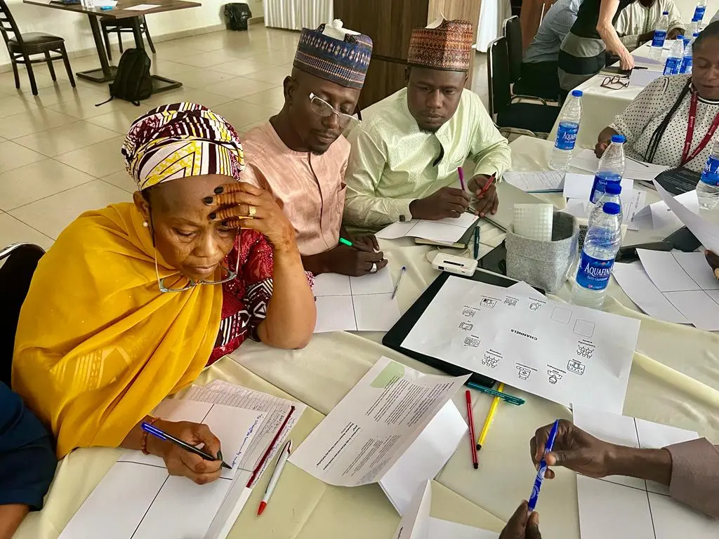 Photograph from a Pathways workshop in Nigeria. 2 men seated at a table look on as a woman in a yellow dress writes on a piece of paper