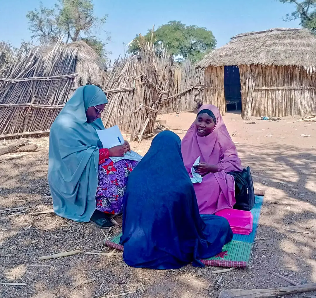 3 women wearing colourful headscarves sit outside a rural home on the ground. Two of the women are holding paper and pens while they speak to a woman in a blue outfit with her back to the camera. 