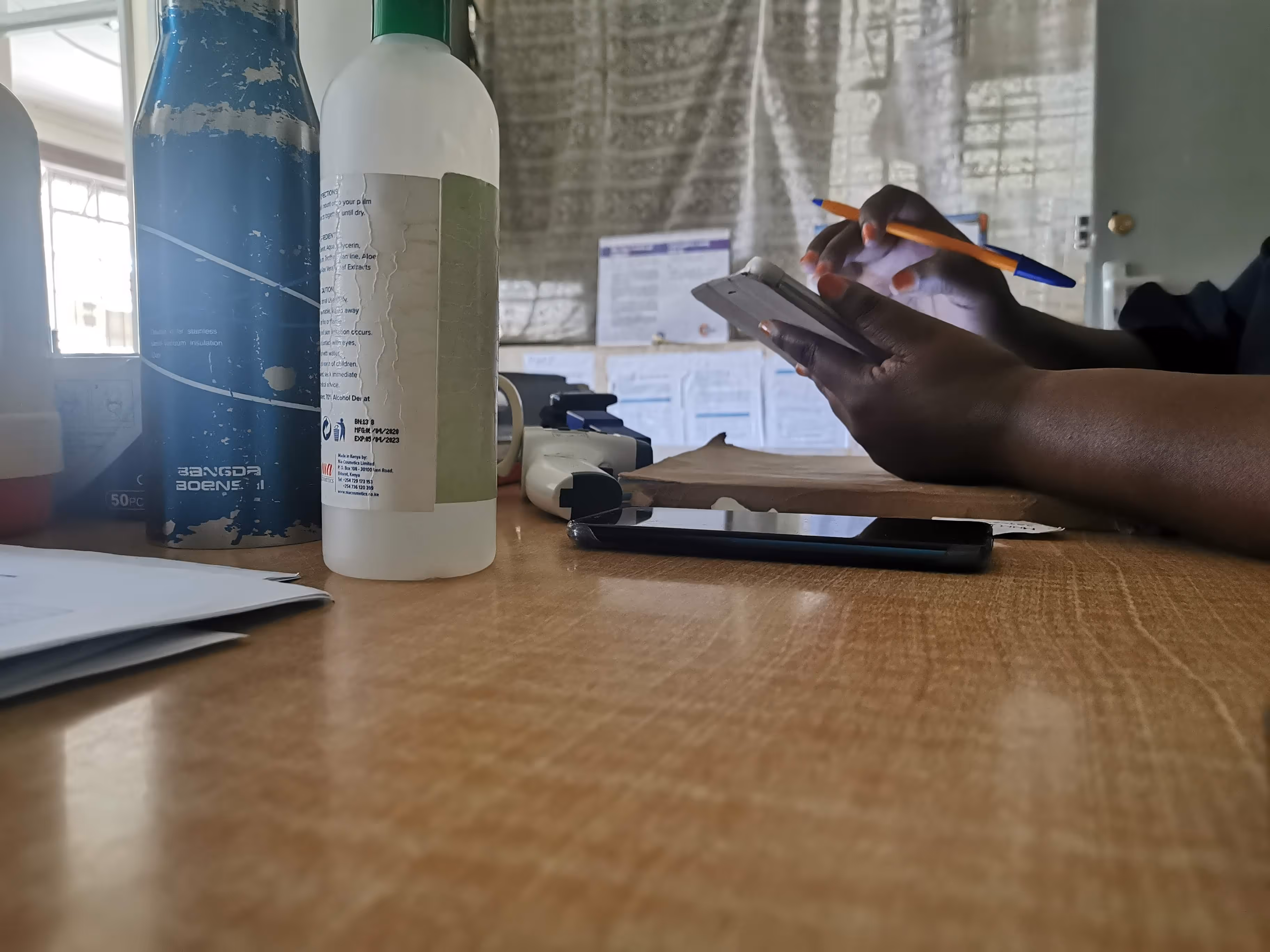 Person writing on a notepad with a pen, sitting at a table with bottles, documents, and a smartphone.