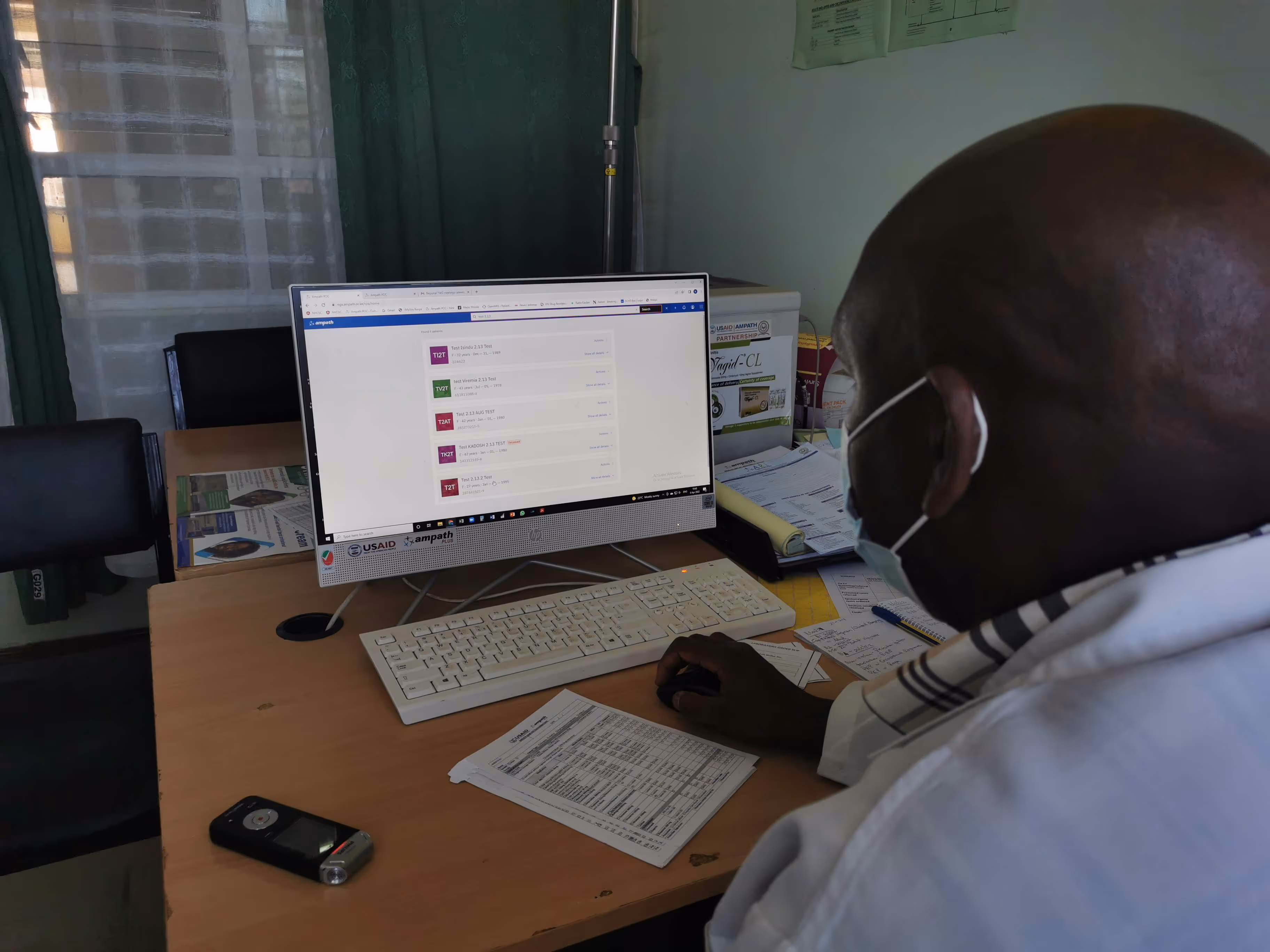 Person wearing a mask working on a desktop computer displaying a list of test entries, seated at a desk with documents and a recorder.