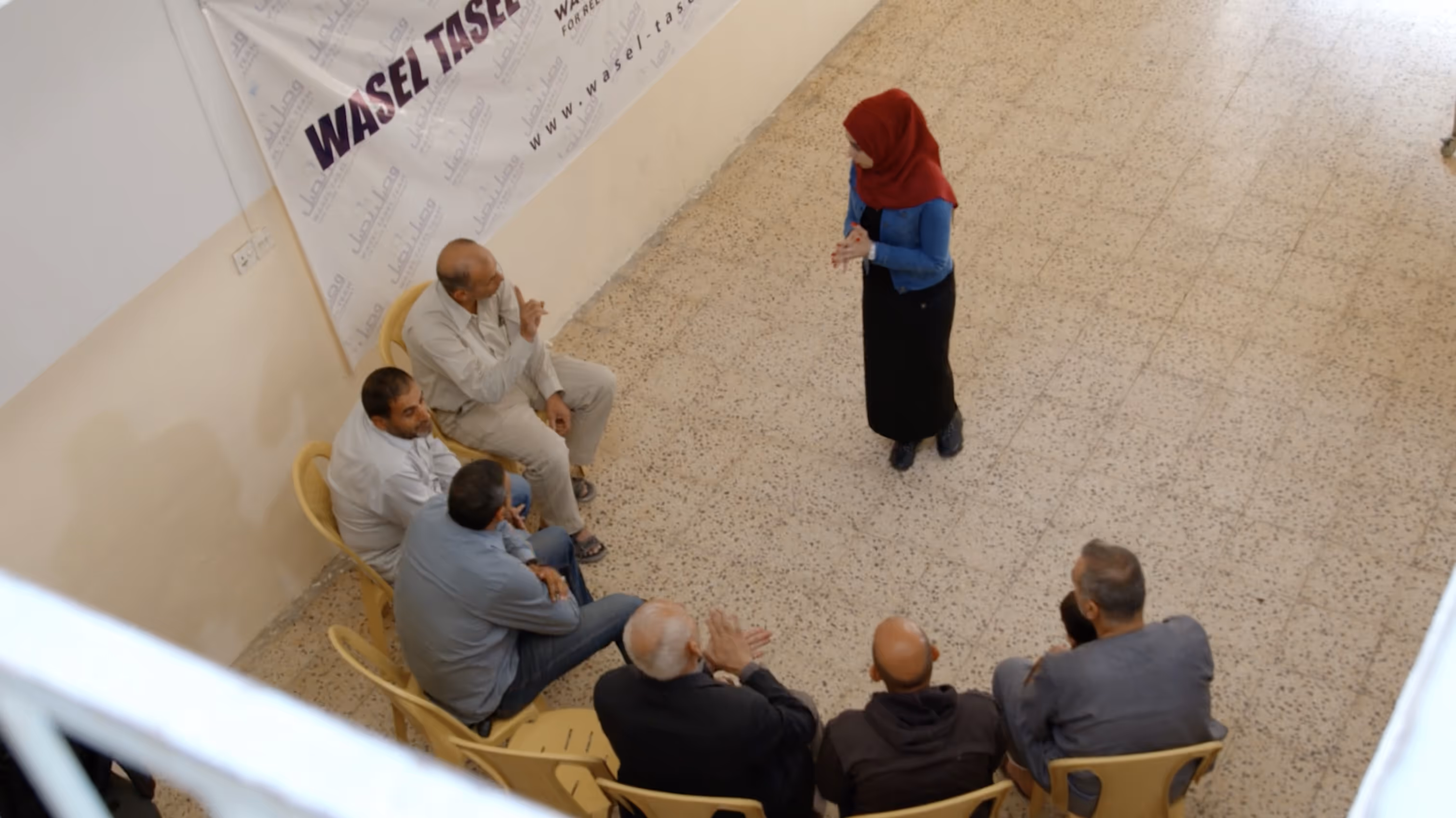 A woman in a red hijab and blue jacket standing and speaking to a seated group of men in yellow chairs in an indoor setting in Mosul, Iraq.