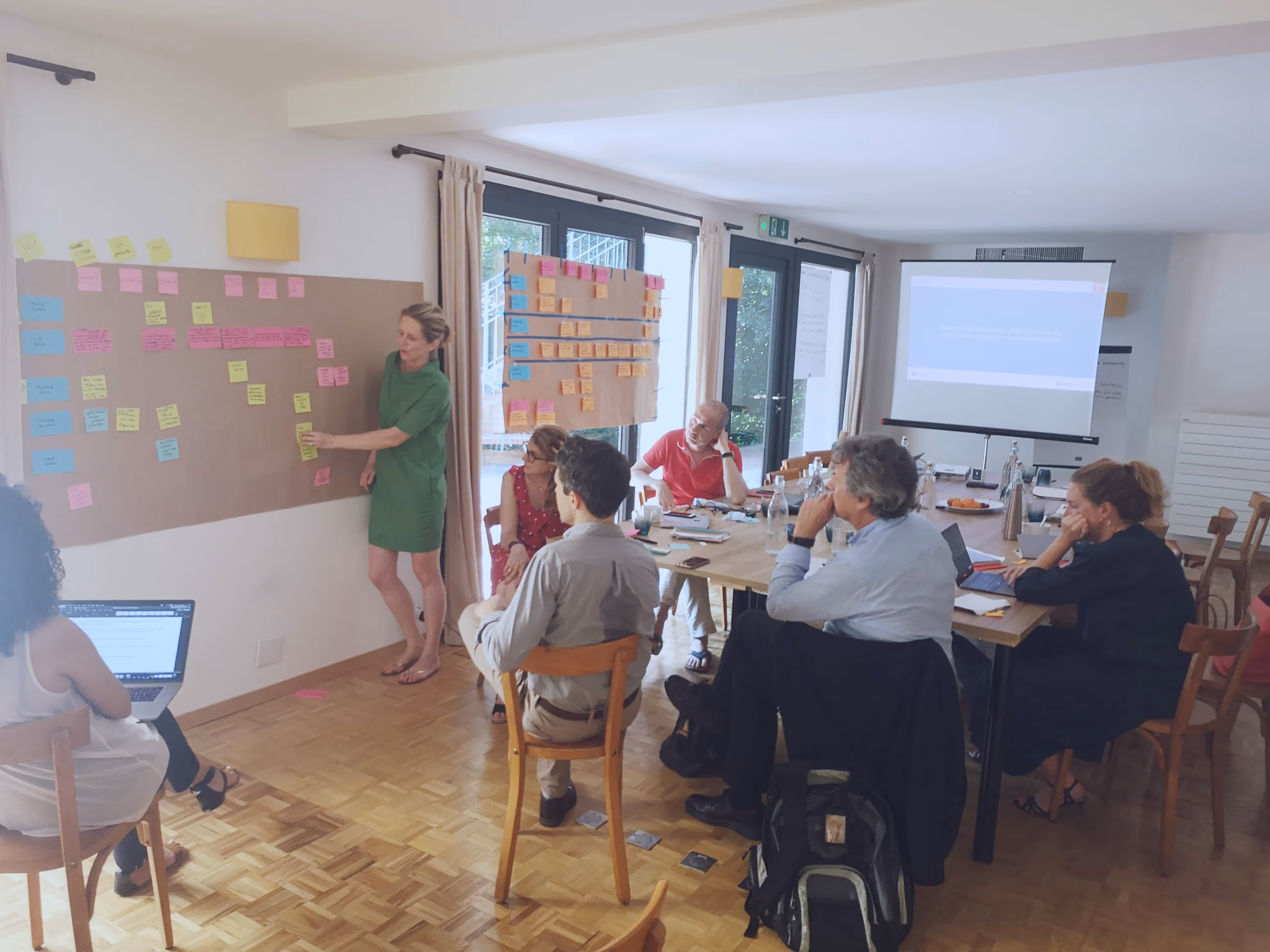 Group of people in a meeting room brainstorming with sticky notes on large sheets of paper on the walls and a projected presentation.