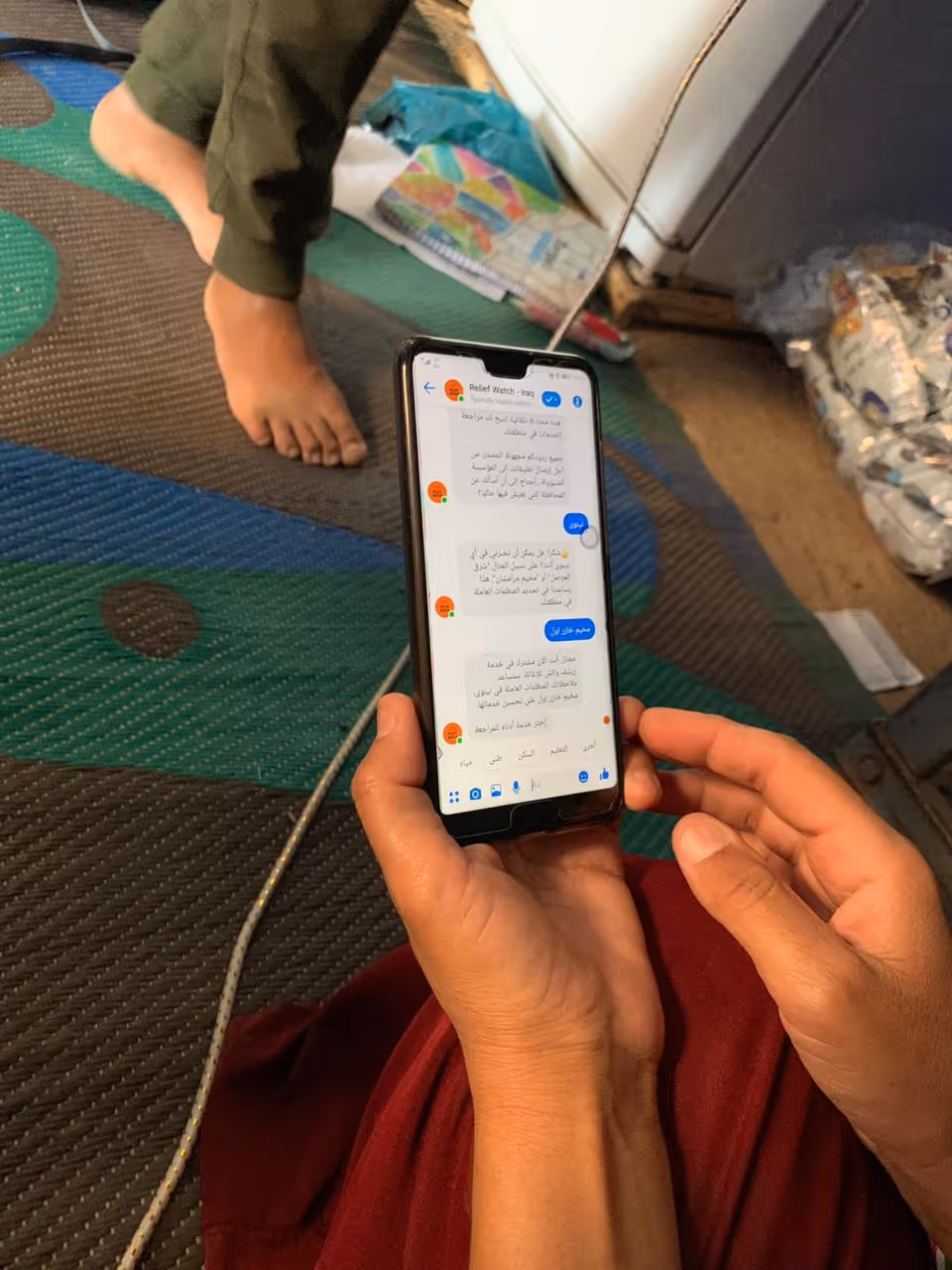 Person holding a smartphone displaying a chat conversation in Arabic with a barefoot in the background on a patterned carpet.