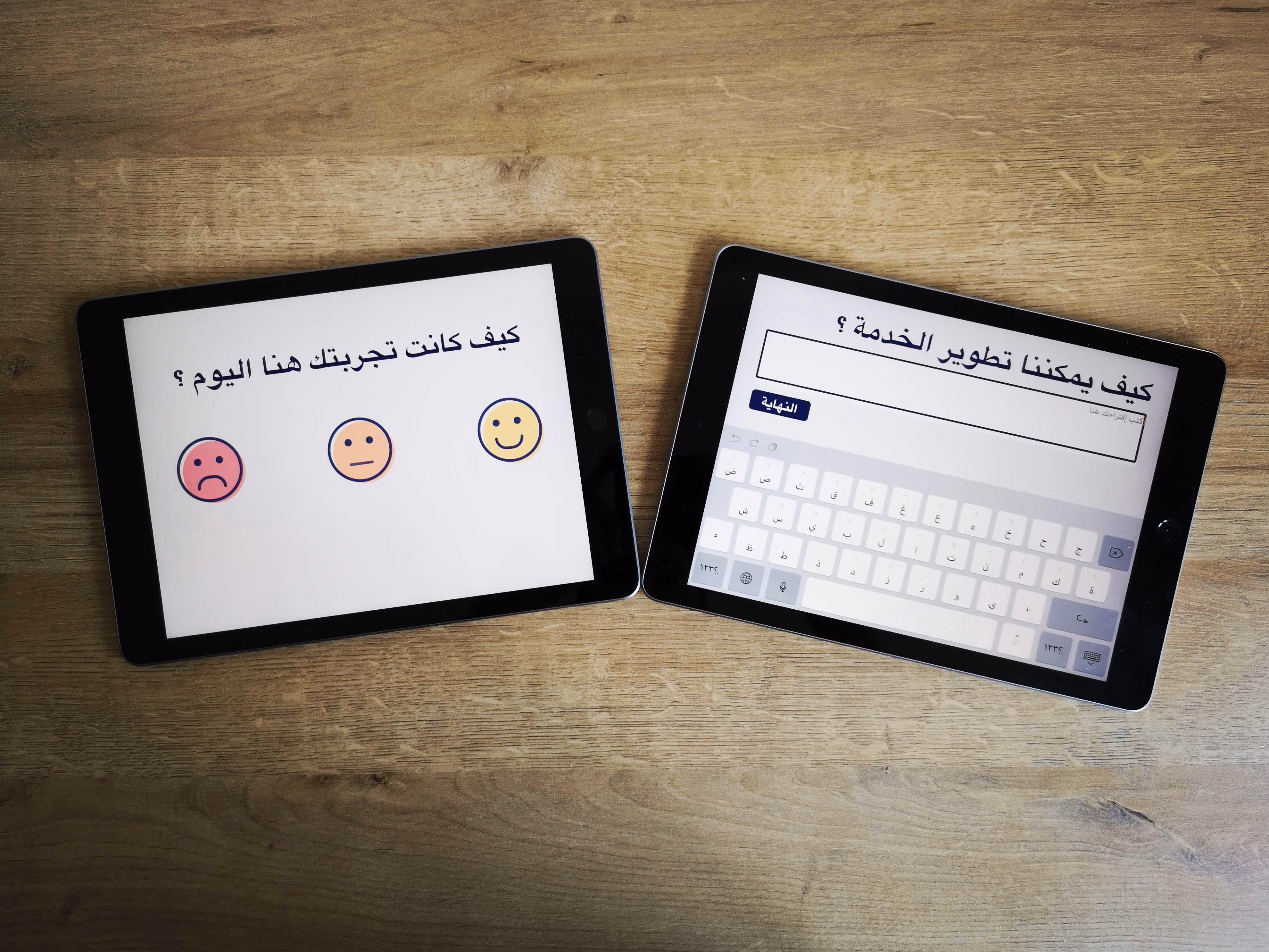 Two tablets on a wooden surface showing Arabic text feedback forms, one with smiley faces for rating experience, the other with a text box and virtual keyboard.