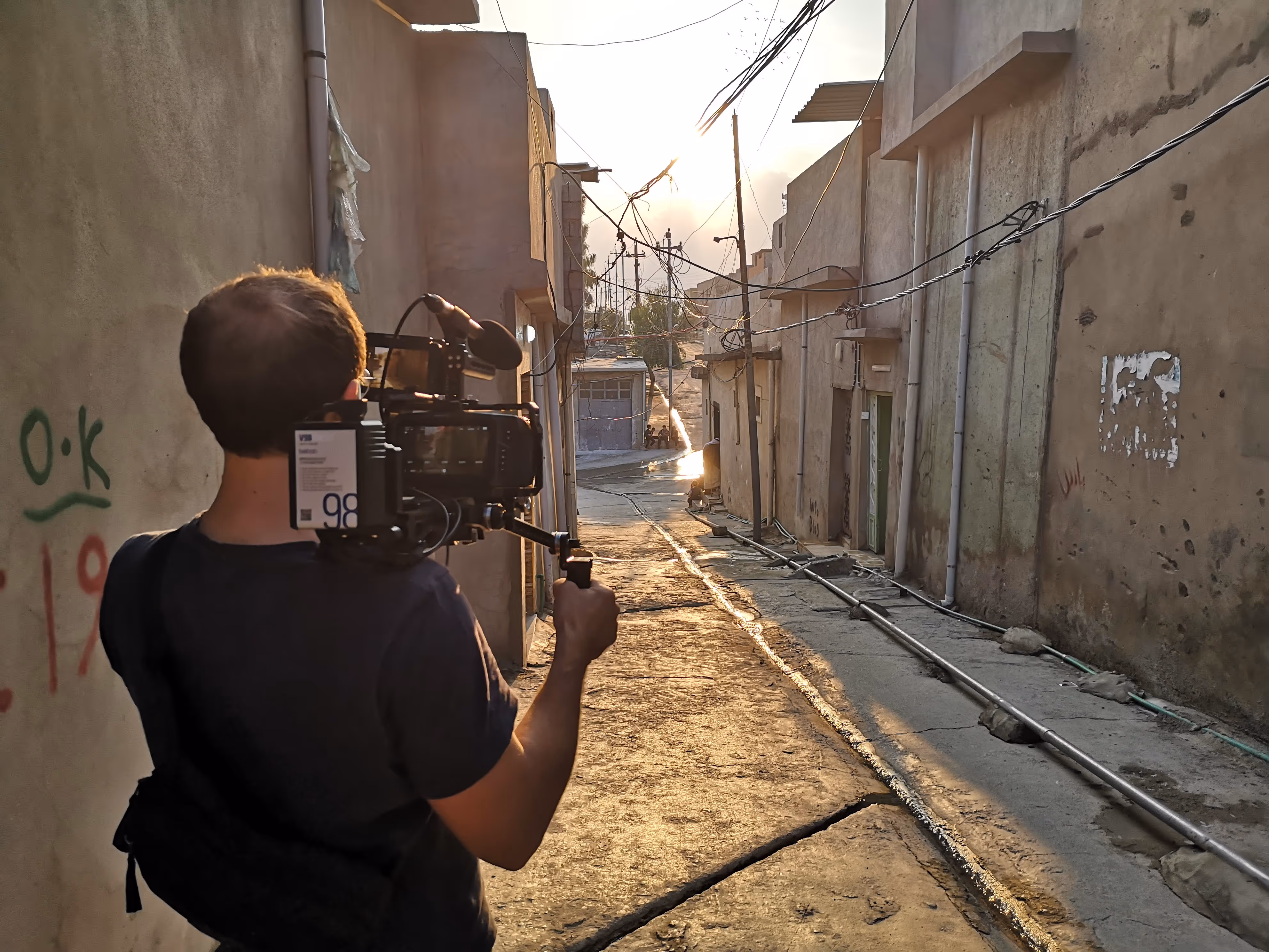 Man holding a video camera filming down a narrow alley at sunset with buildings on both sides.