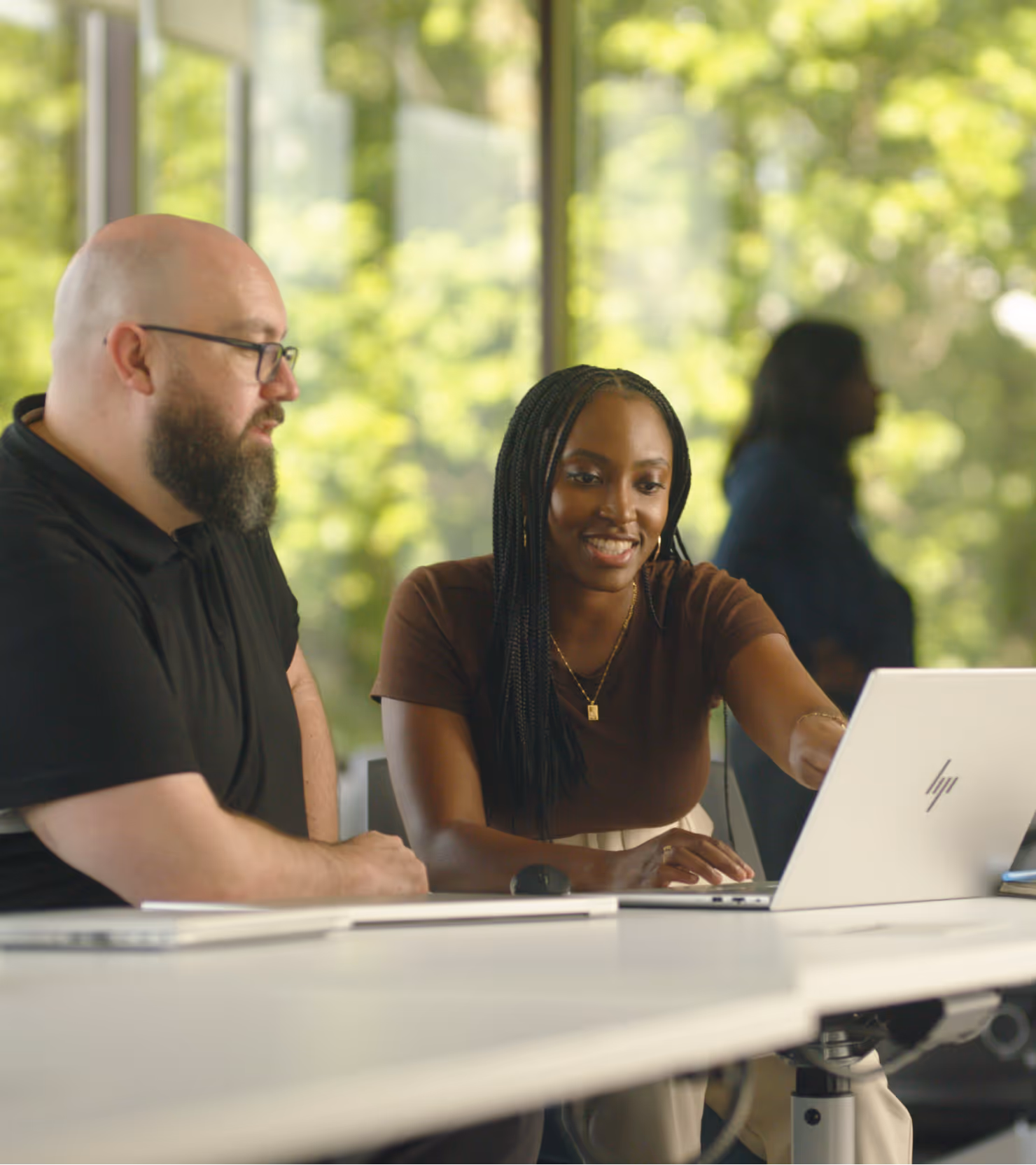 A woman and a man sitting at a desk, looking at a laptop screen with a green outdoor background.