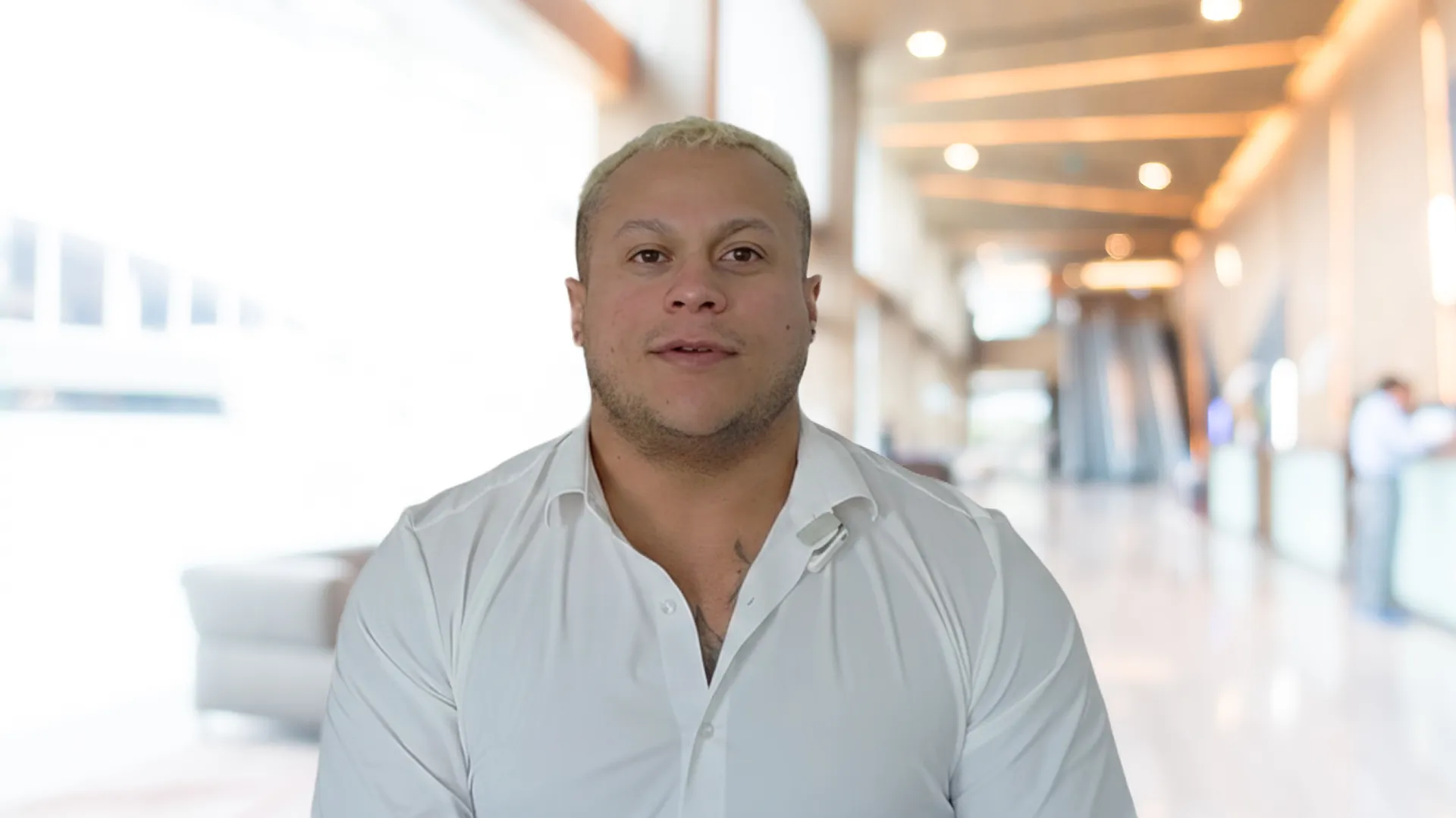 Man with short blond hair wearing a white shirt, standing in a bright modern lobby with escalators and people in the background.