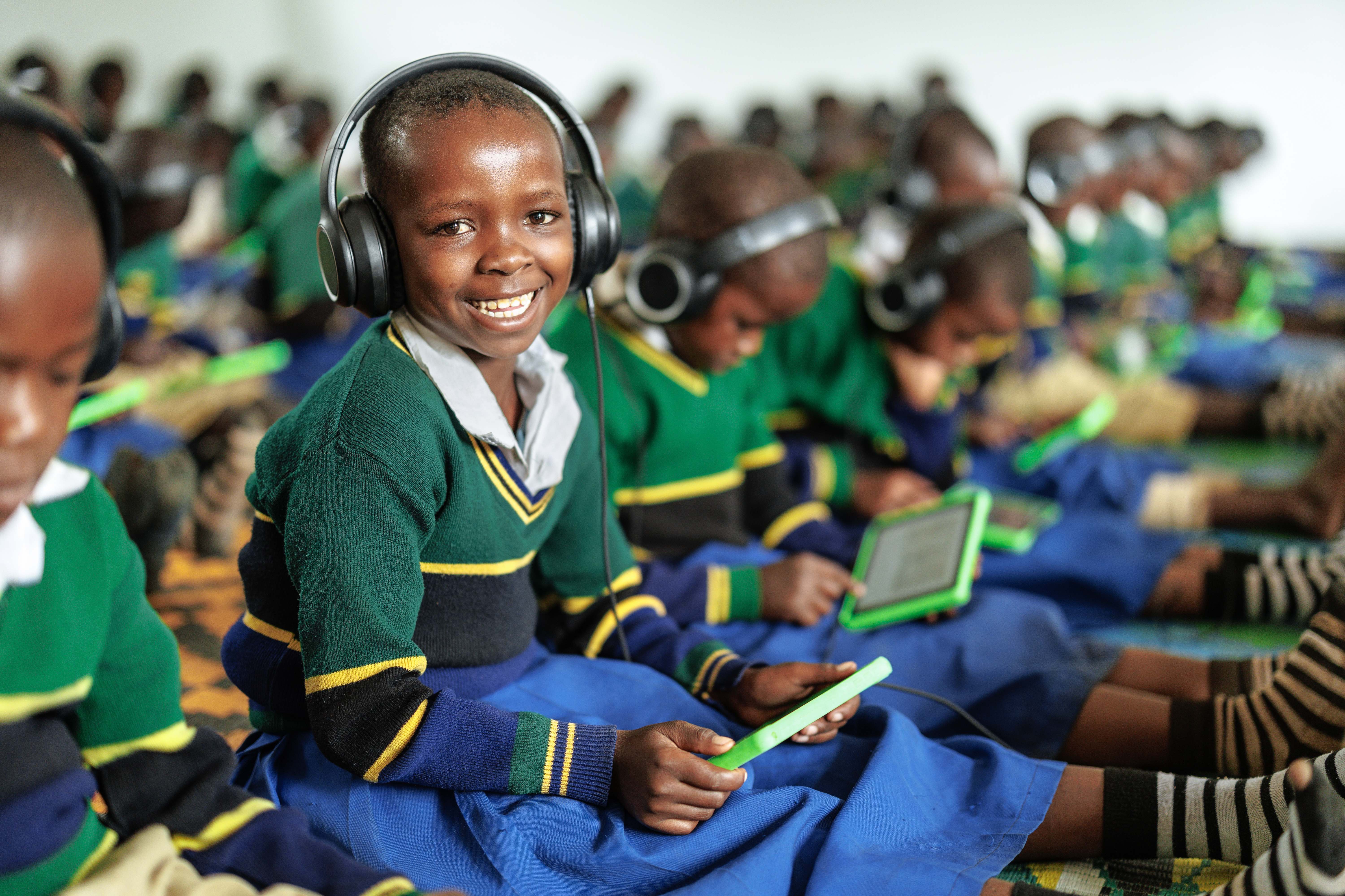 A smiling child wearing headphones holds a tablet in a classroom. Many children in uniform, focused on tablets, create a lively, engaging atmosphere.