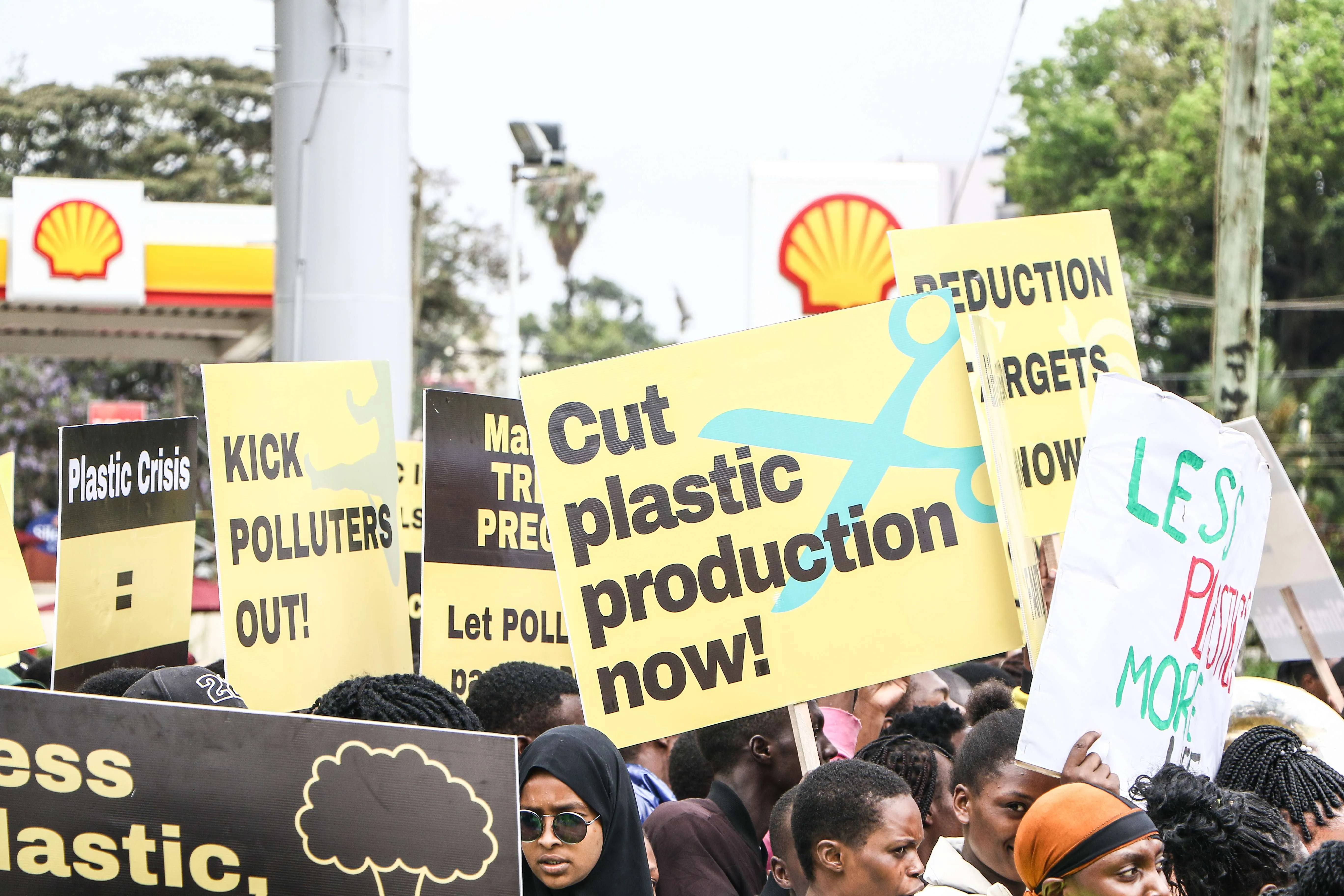 Protesters hold signs with anti-plastic messages like "Cut plastic production now!" and "Kick polluters out!" near a Shell gas station, conveying urgency.