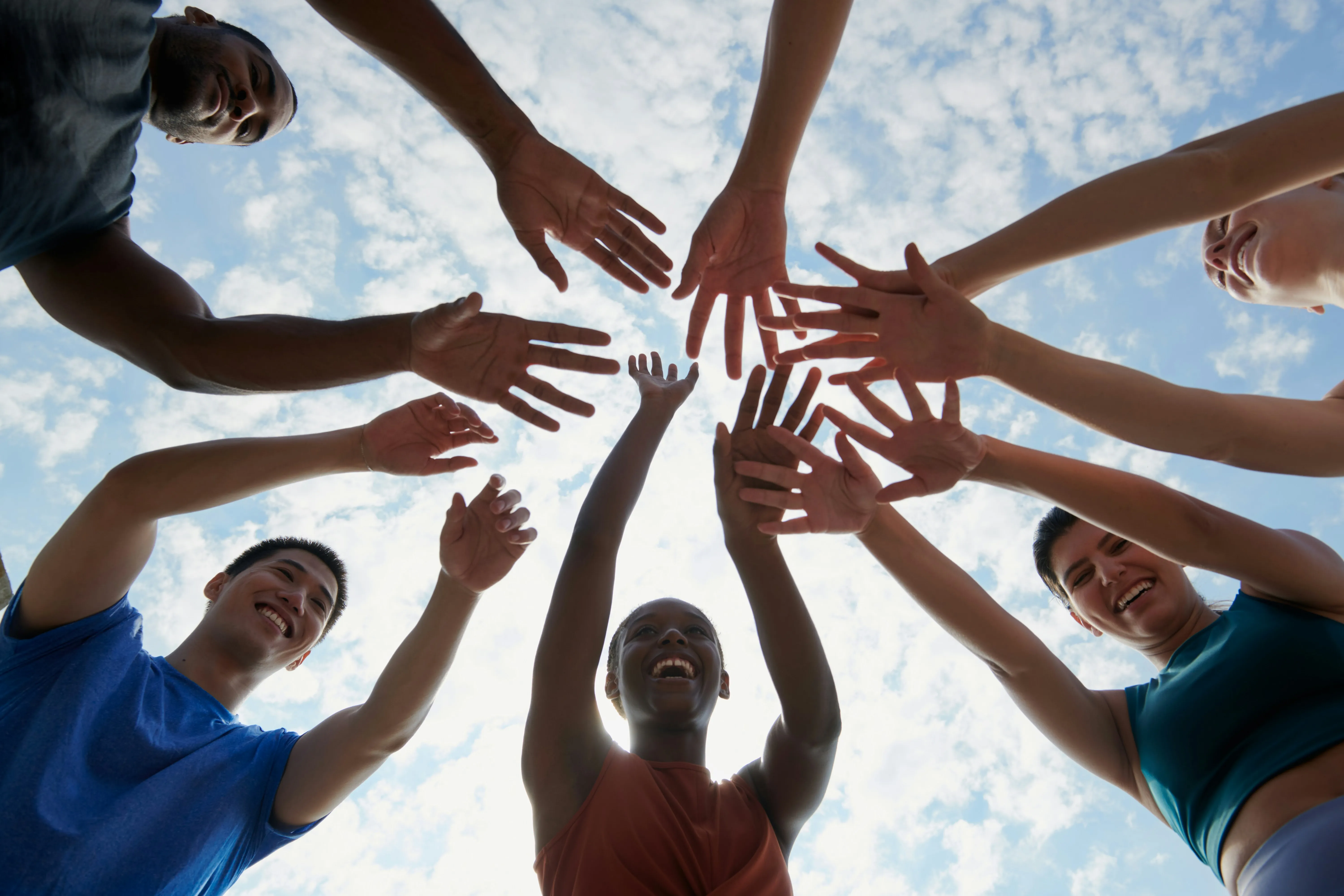 A group of people standing in a circle, smiling, and reaching their hands into the center against a blue sky with clouds, conveying unity and joy.