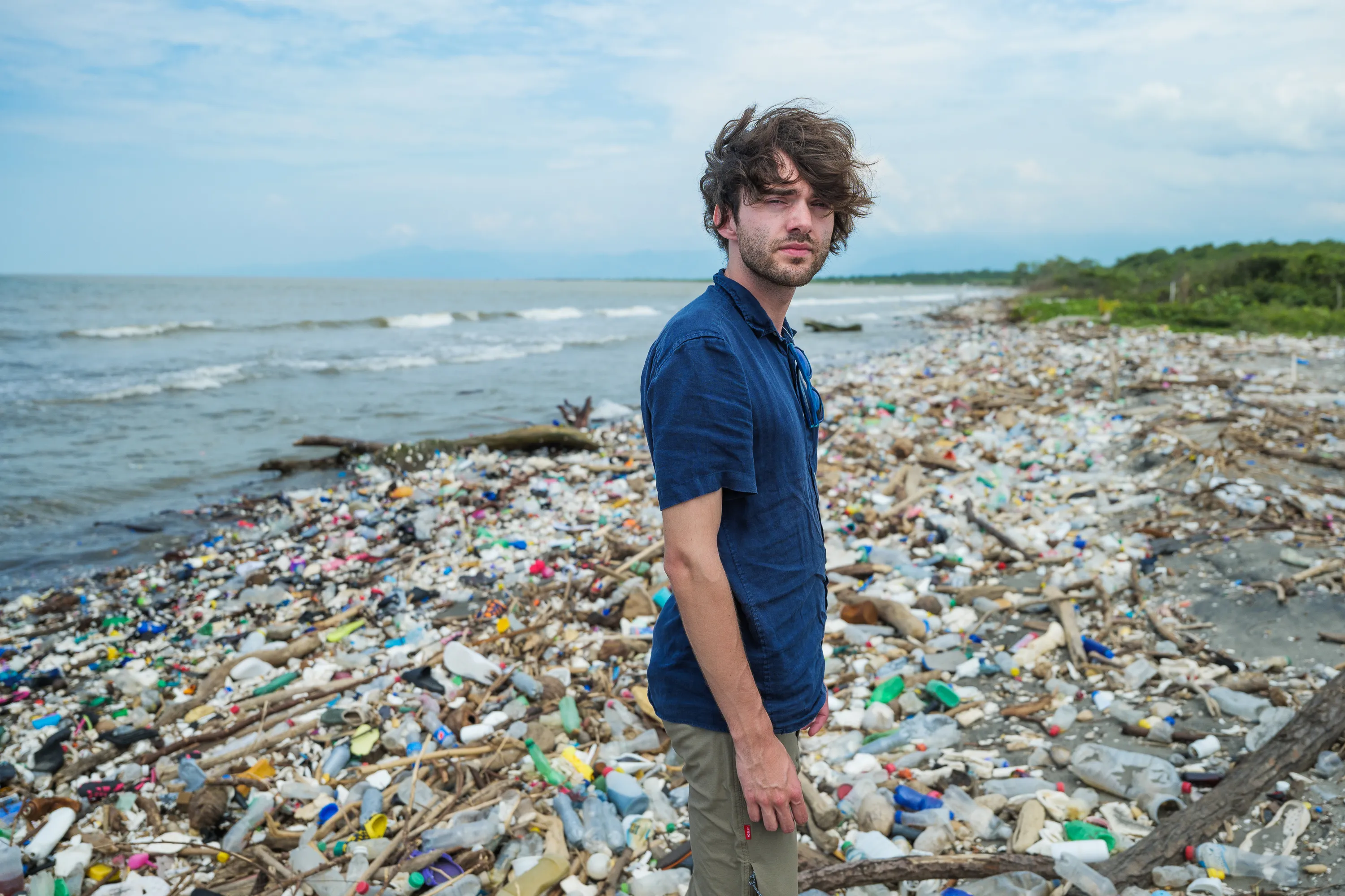 A person in a blue shirt stands on a heavily polluted beach, covered in plastic debris and trash. The expression is serious, reflecting environmental concern. 