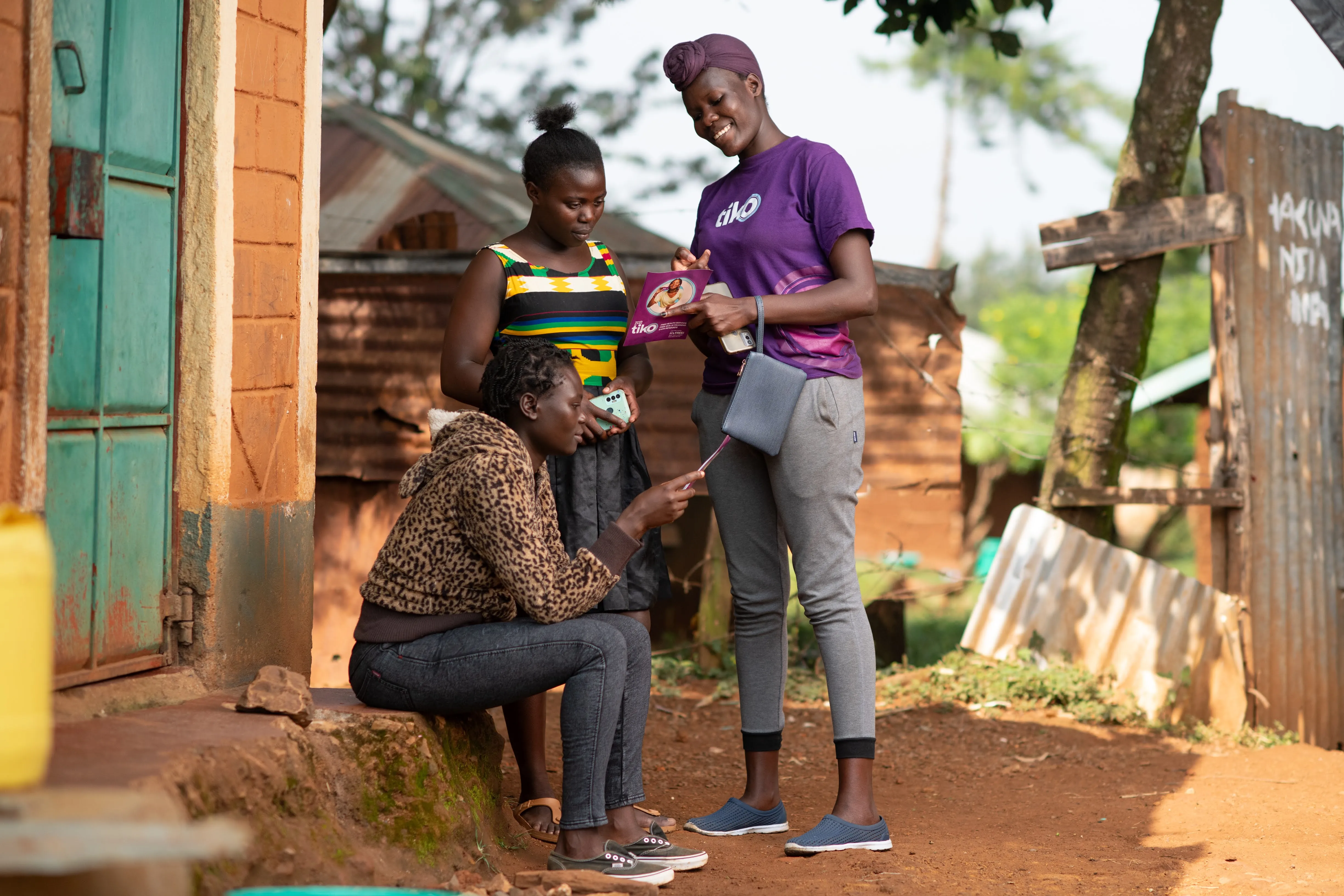 Three people are gathered outside near a brick building. One is seated, pointing at a Tiko pamphlet held by another person, who is smiling. The mood is engaged and friendly.