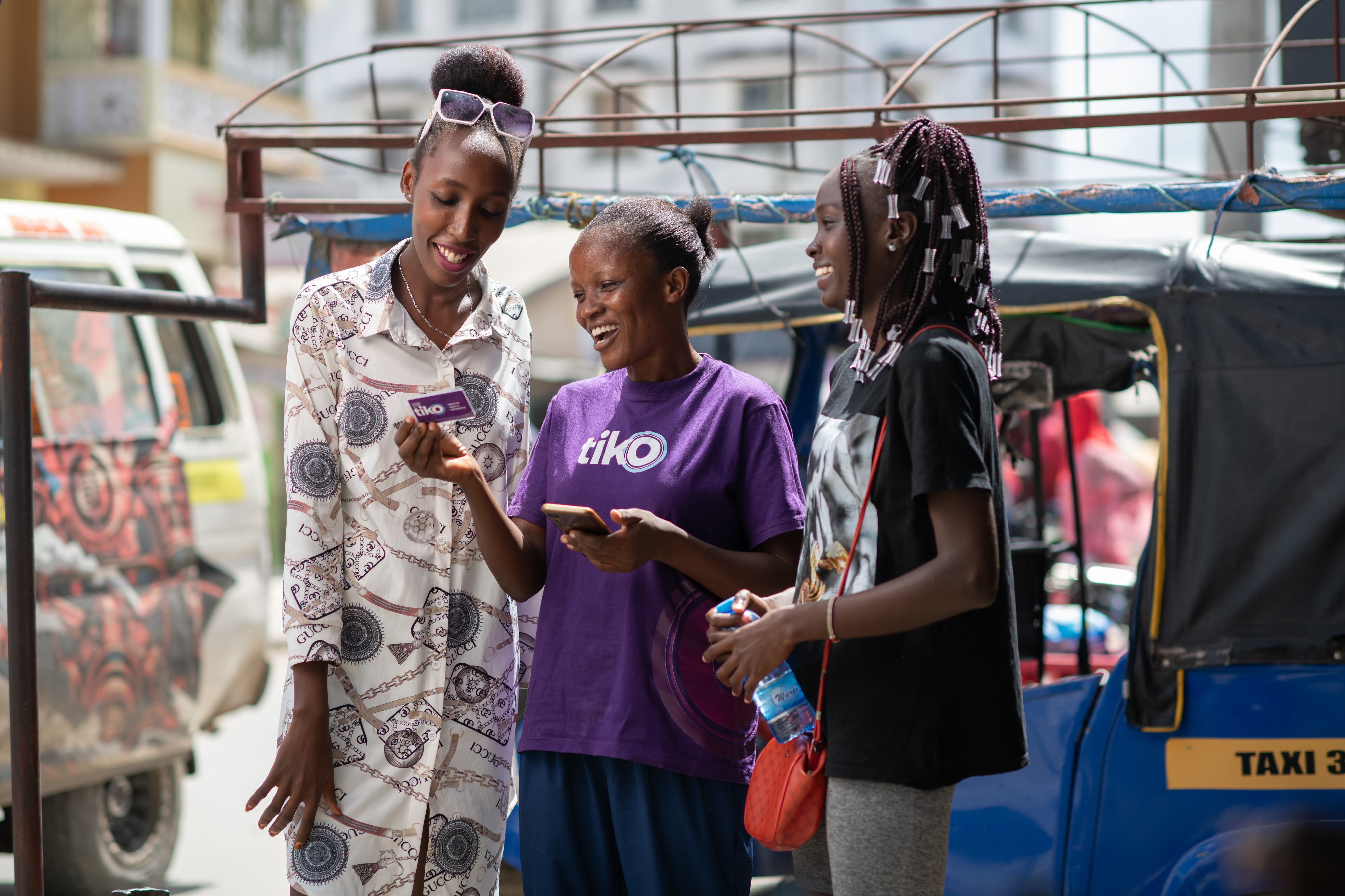 Three people are gathered outside near a brick building. Looking at Tiko pamphlet held by another person, who is smiling. The mood is engaged and friendly.